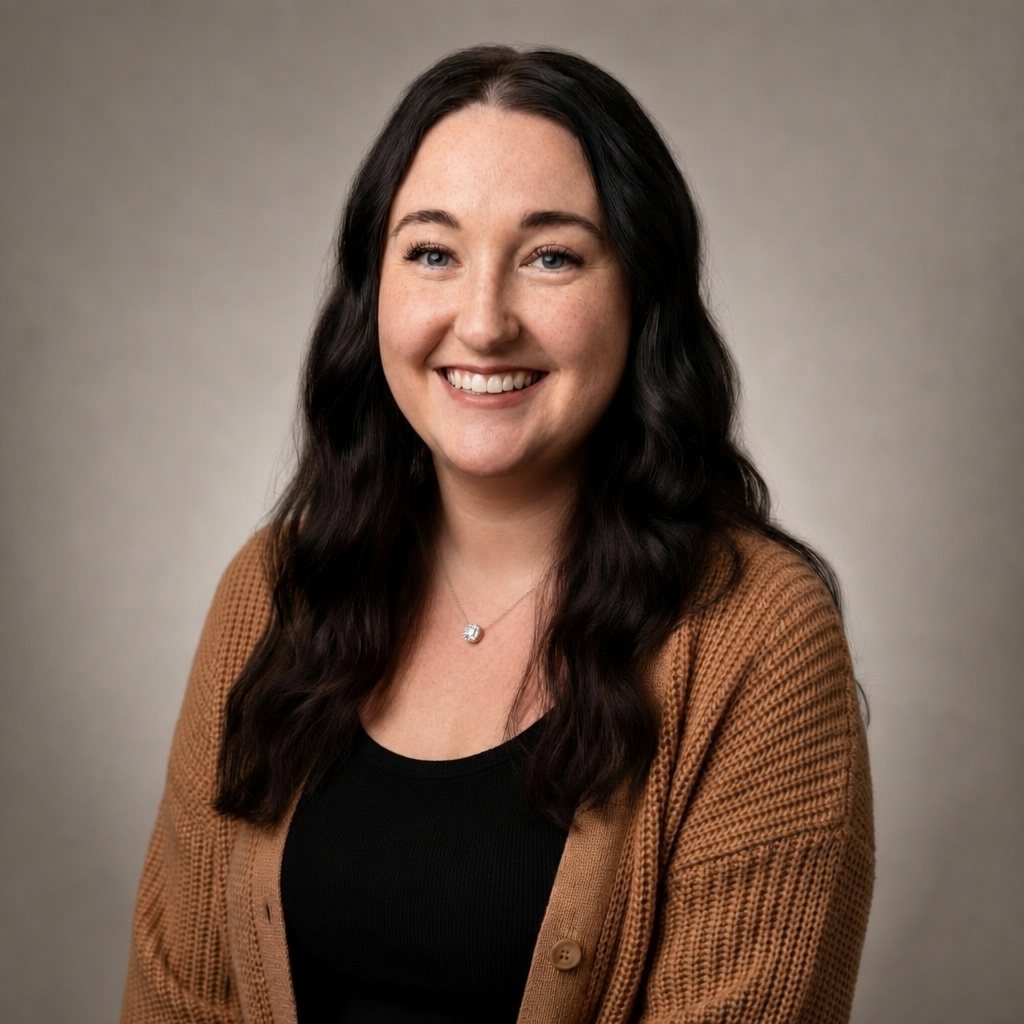 A woman with long, dark wavy hair, blue eyes, and freckles, smiling and wearing a black top, a brown cardigan, and a silver necklace in front of a plain, neutral background.