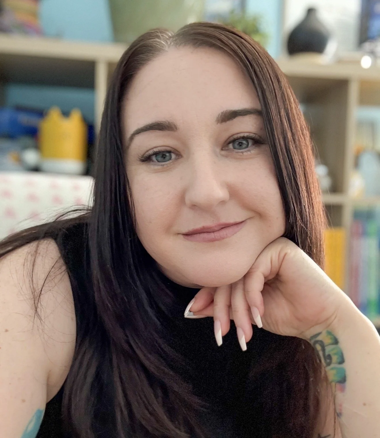 Close-up of a woman with long dark hair and blue eyes, resting her chin on her hand, in an indoor setting with shelves and decorative objects in the background.