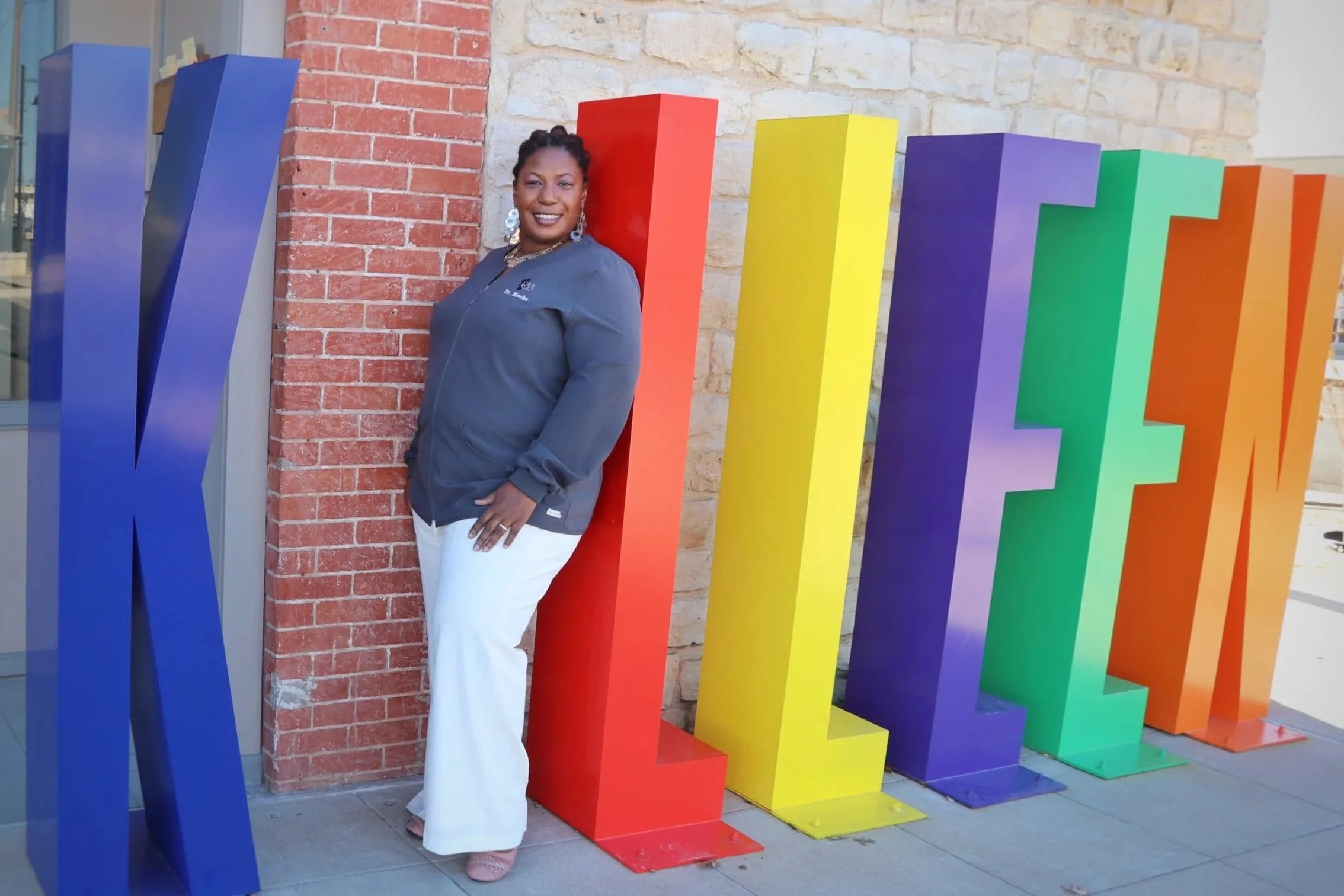 Dr. Iperlitta "Mecka" Lolis leaning against a red letter on a colorful display spelling Killeen in rainbow colors outside a building with a brick wall.