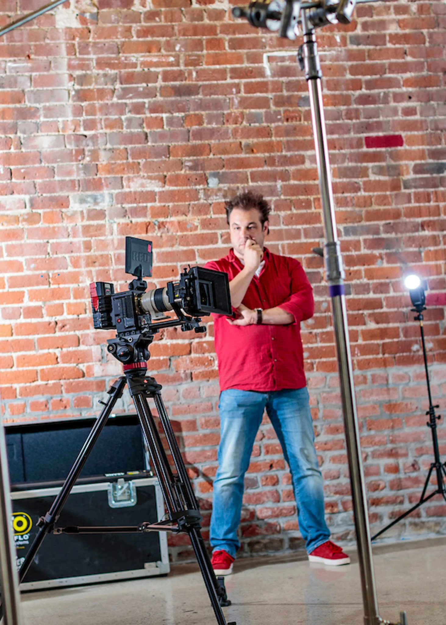 A man in a red shirt and jeans standing behind a professional camera on a tripod, observing and thinking during a video shoot in front of a brick wall.