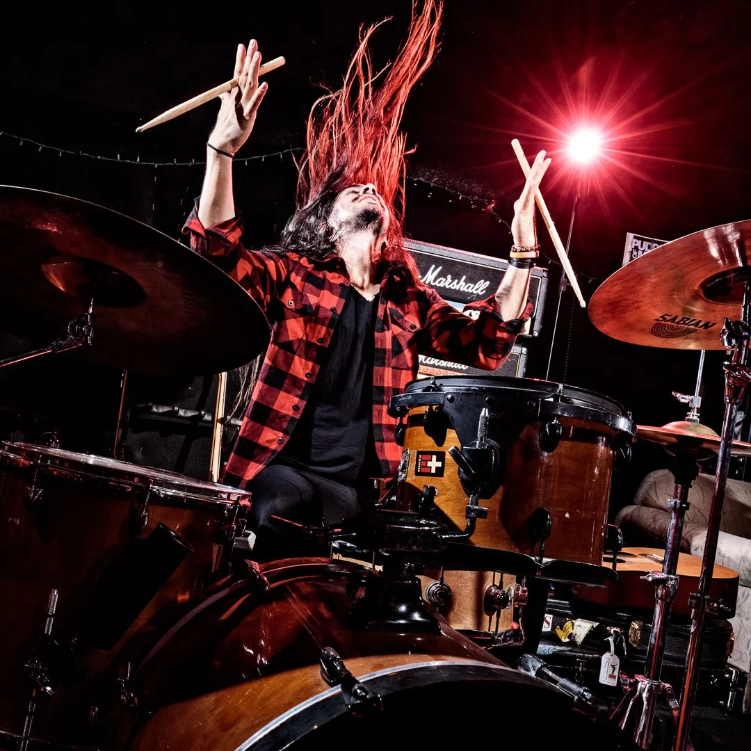 A drummer with long hair, wearing a red and black plaid shirt, is playing drums passionately with their hair flying in the air. The background includes a Marshall amplifier and stage lights, with one bright red stage light shining.
