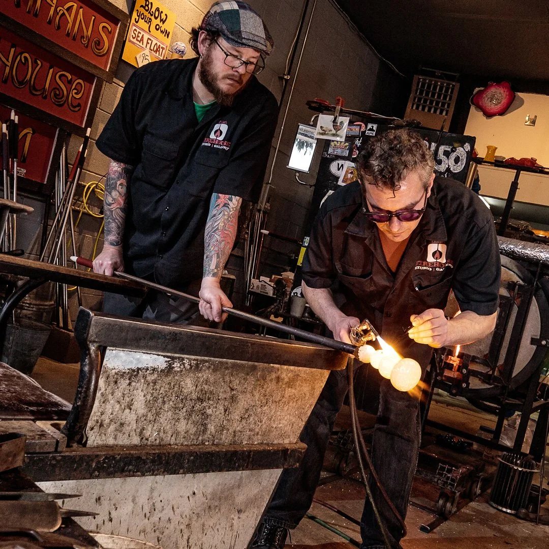 Two men working in a blacksmith shop, one is heating metal with a torch while the other observes. The man with the torch is wearing sunglasses, and the other has tattoos and wears glasses and a cap. The shop has signs and tools related to blacksmithing.
