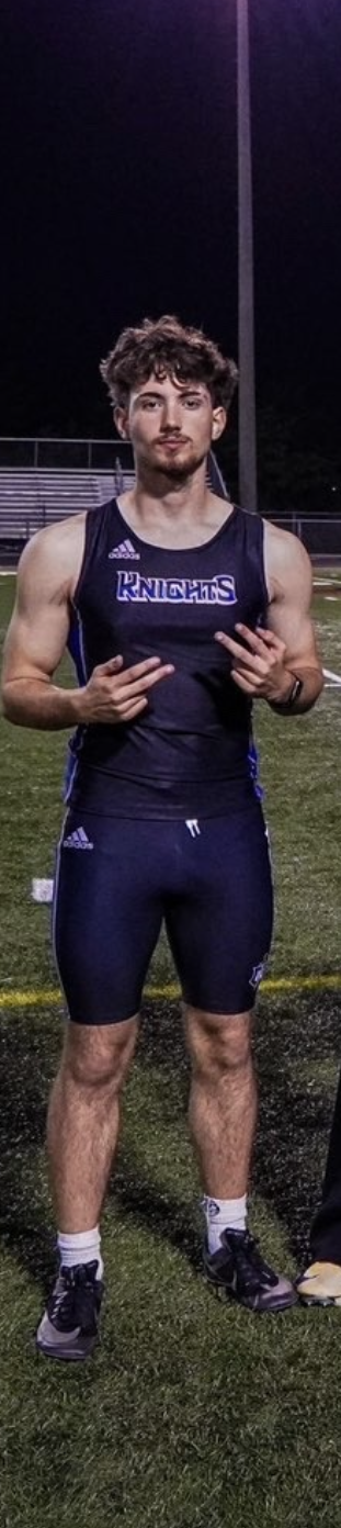 Young male athlete standing on a football field at night, wearing a navy blue sports uniform with 'KNIGHTS' written on the chest, holding up peace signs with both hands.
