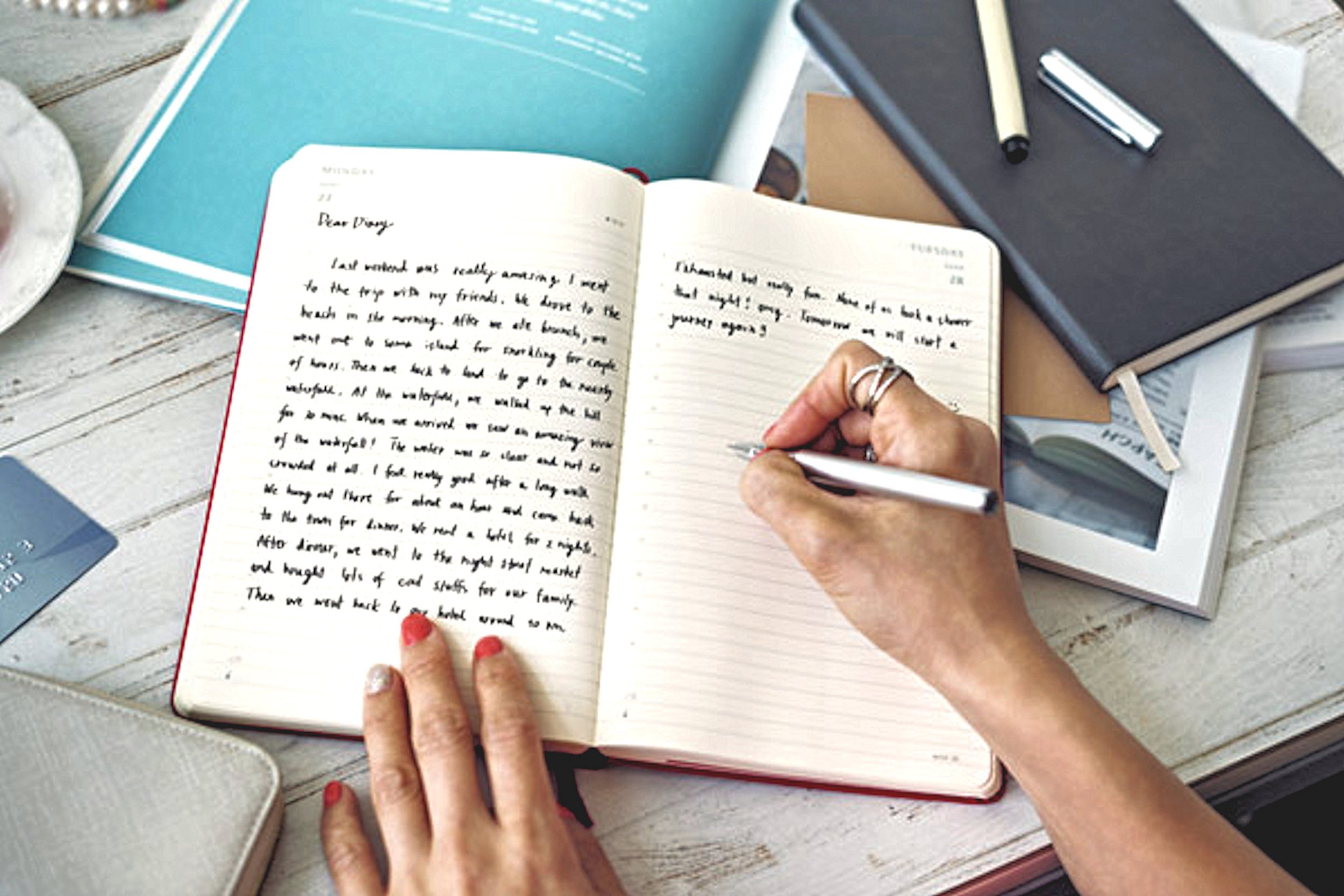 A person with painted nails is writing in a notebook with a pen. The notebook is open on a wooden surface surrounded by other notebooks and pens.