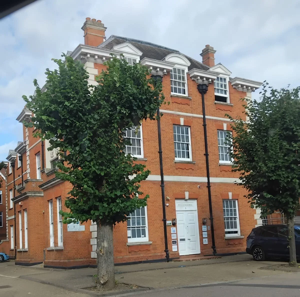 A brick building with white-framed windows and decorative architectural features, flanked by trees and parked cars on the street.