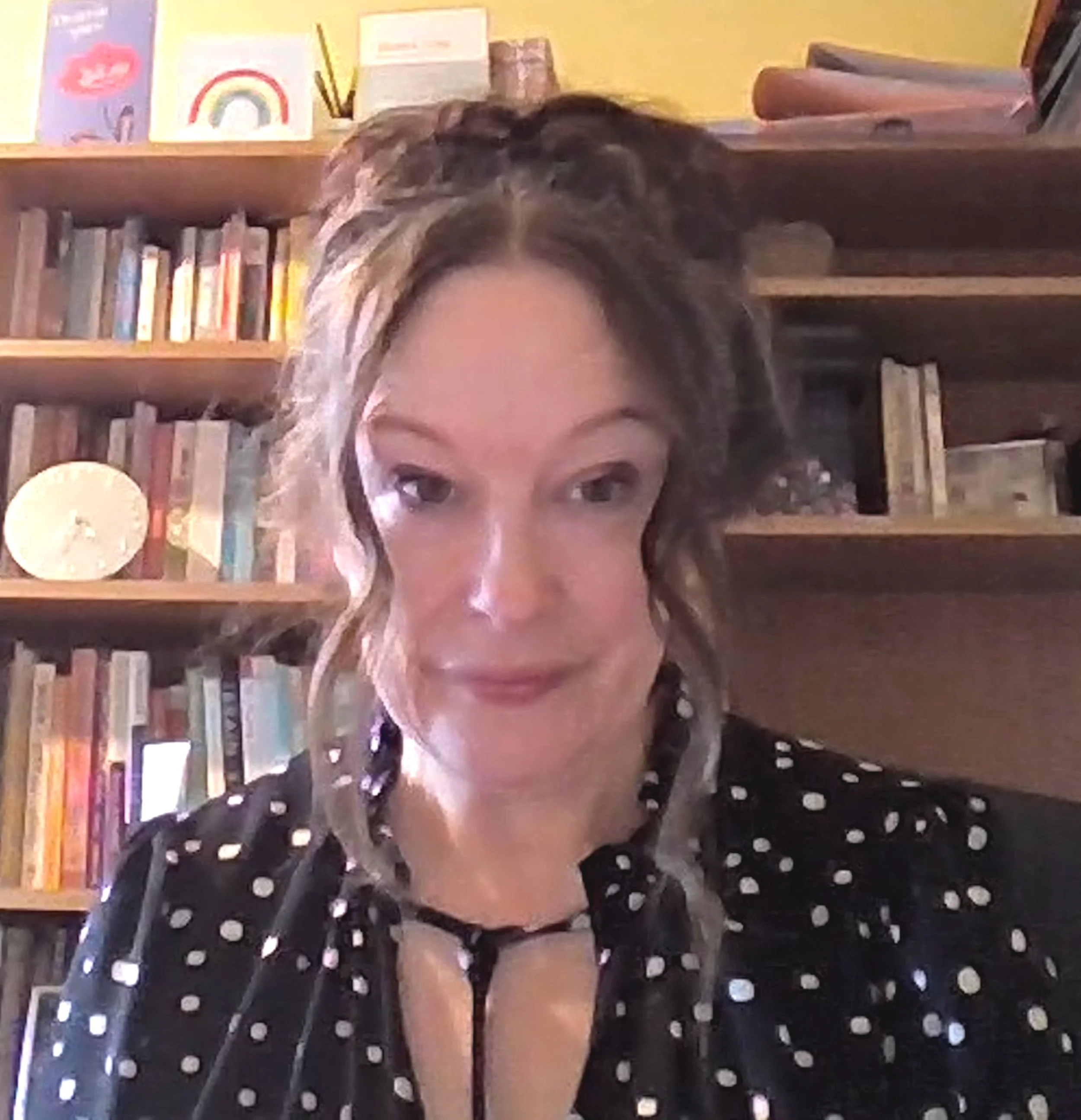 A person with curly hair in front of a bookshelf, wearing a black polka dot top and black lace underwear, with the top part of their face showing.