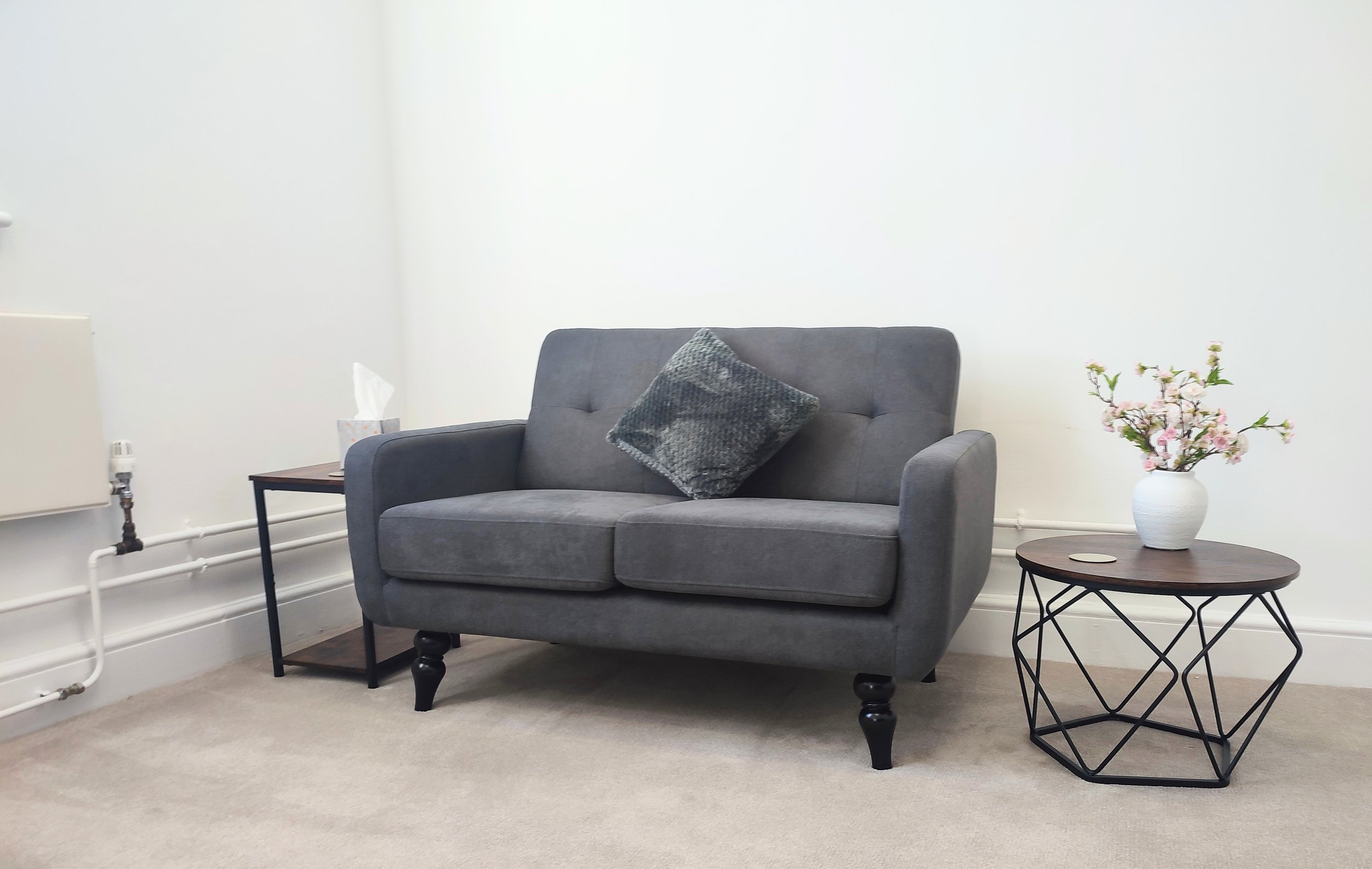 A living room with a gray sofa, black wooden legs, a decorative pillow, a side table with a tissue box, a white vase with pink and white flowers, and a round coffee table with a wireless charging pad, against a white wall with a radiator and pipes.