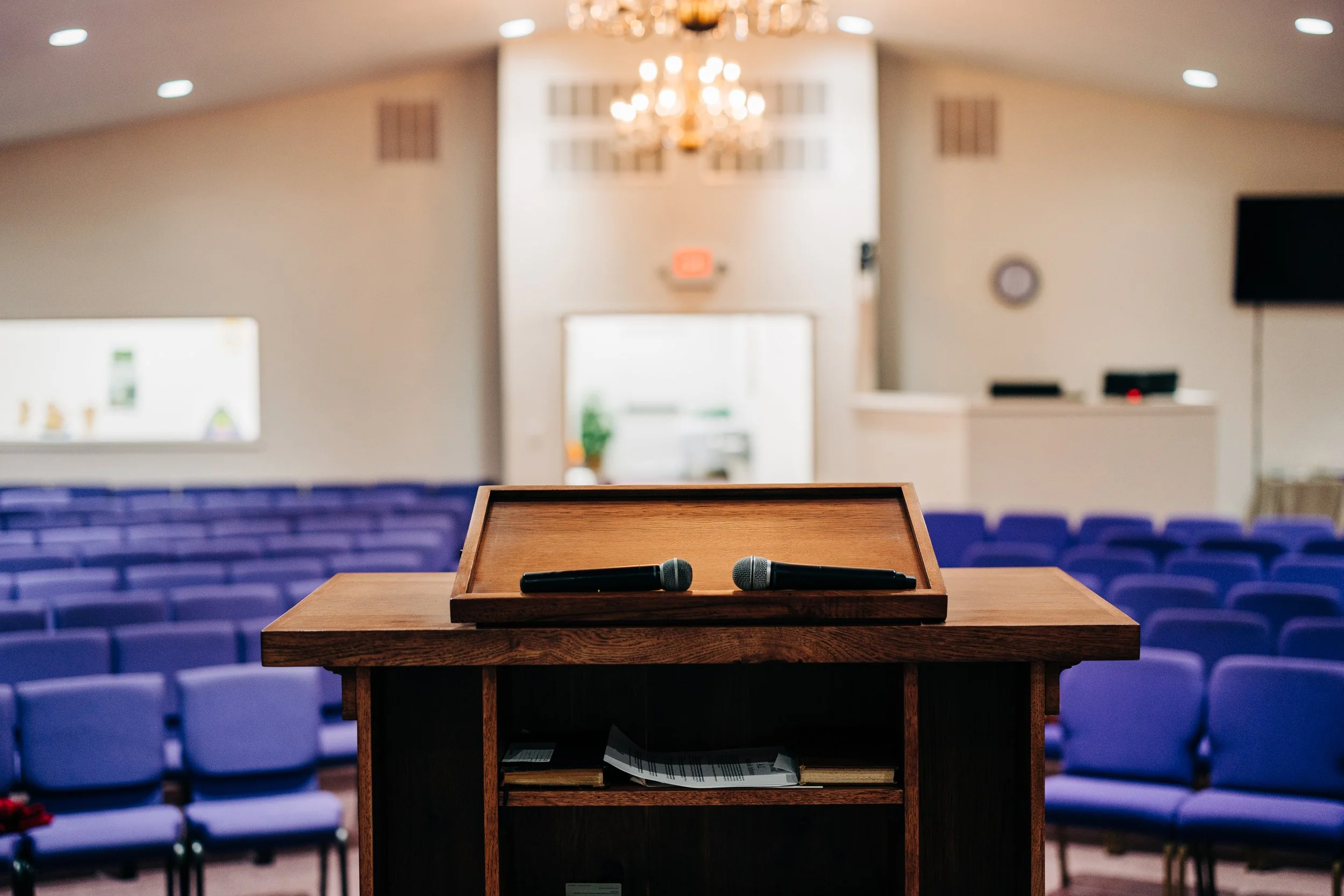 Wooden podium with two microphones in an empty church auditorium with blue seating and a chandelier