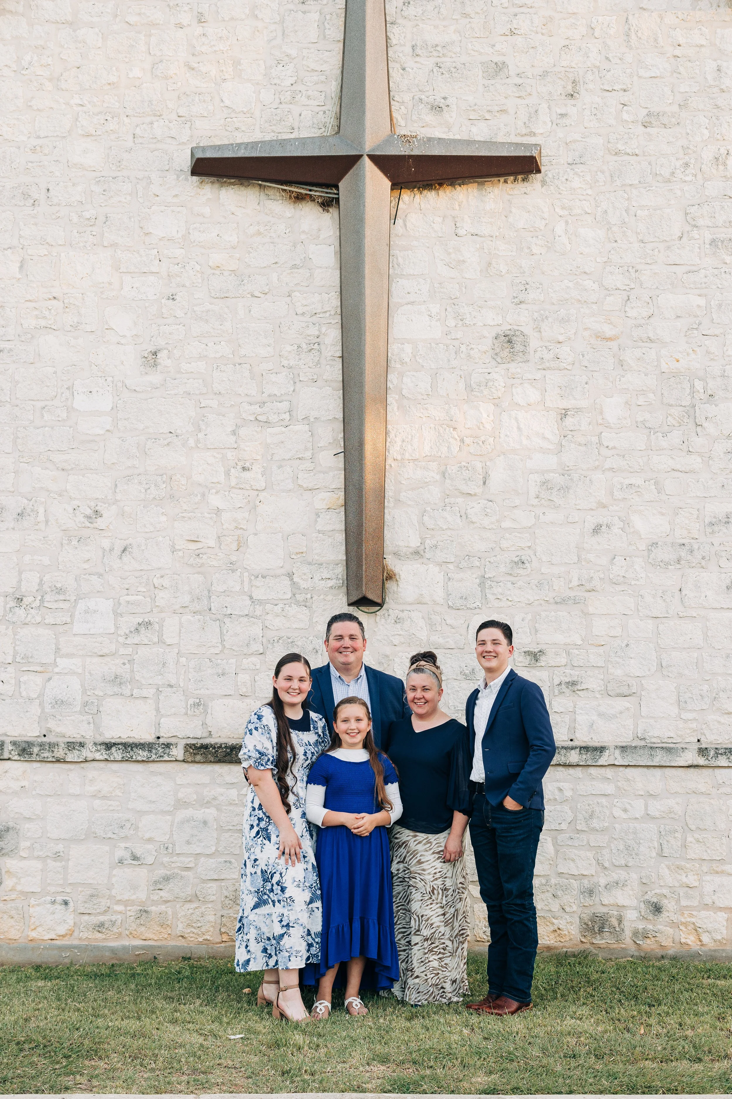 Family posing beneath a large metal cross on a white stone church wall
