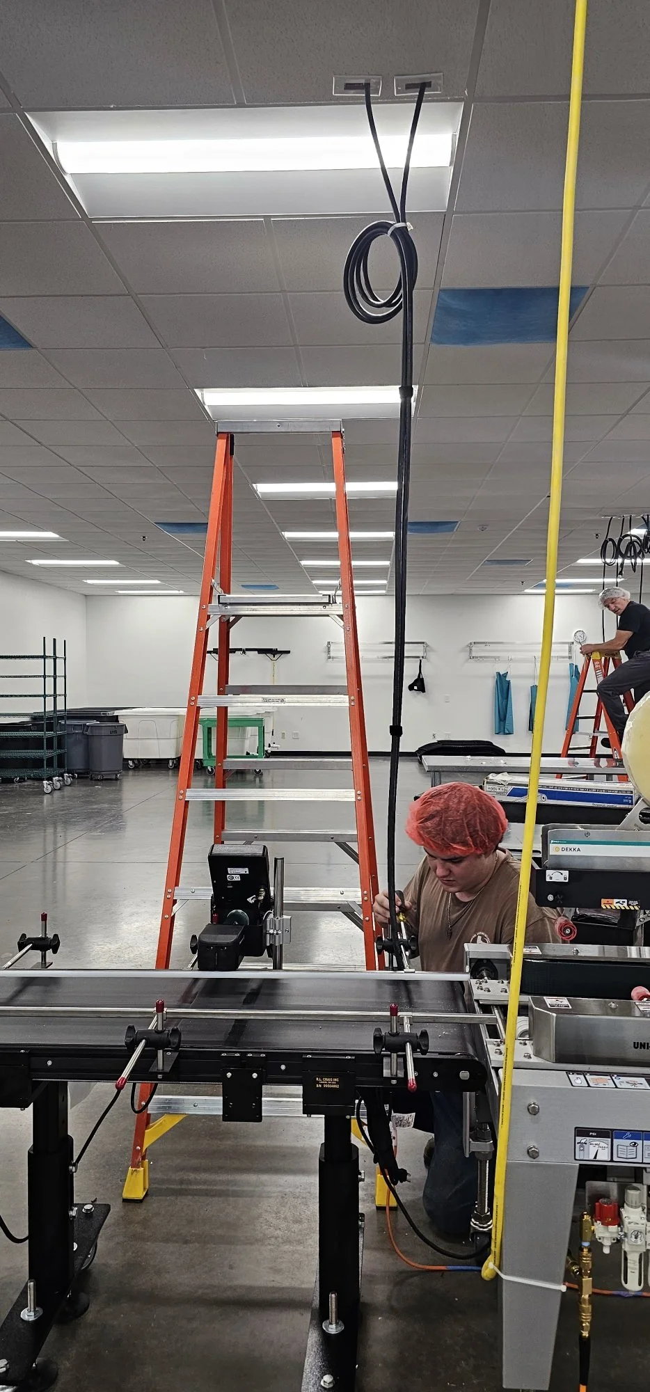 A worker in a hairnet using a machine in a large industrial room with a ladder, overhead lighting, and various equipment.
