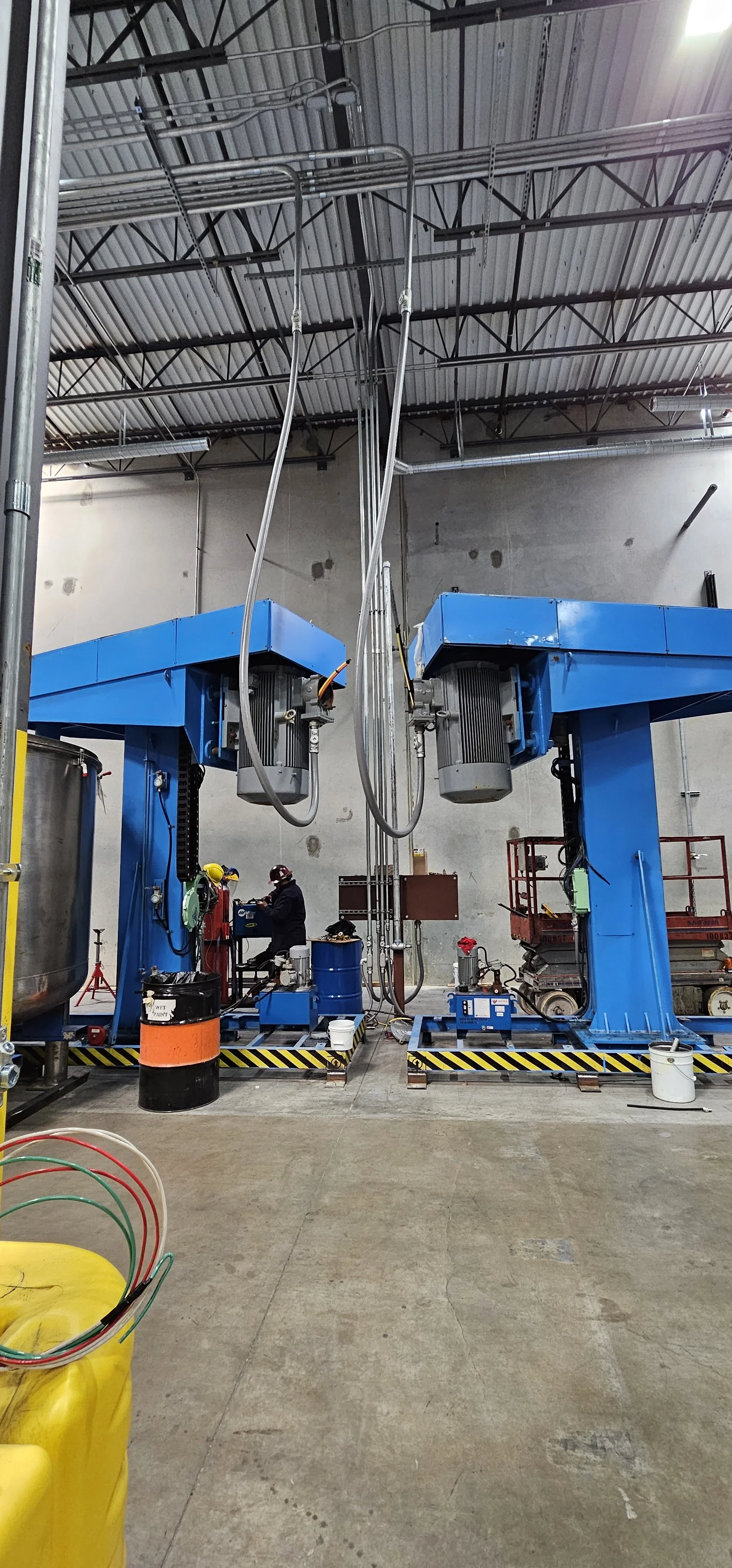 Industrial workshop with two large blue hydraulic lifts, a worker in safety gear, various equipment, and construction supplies.