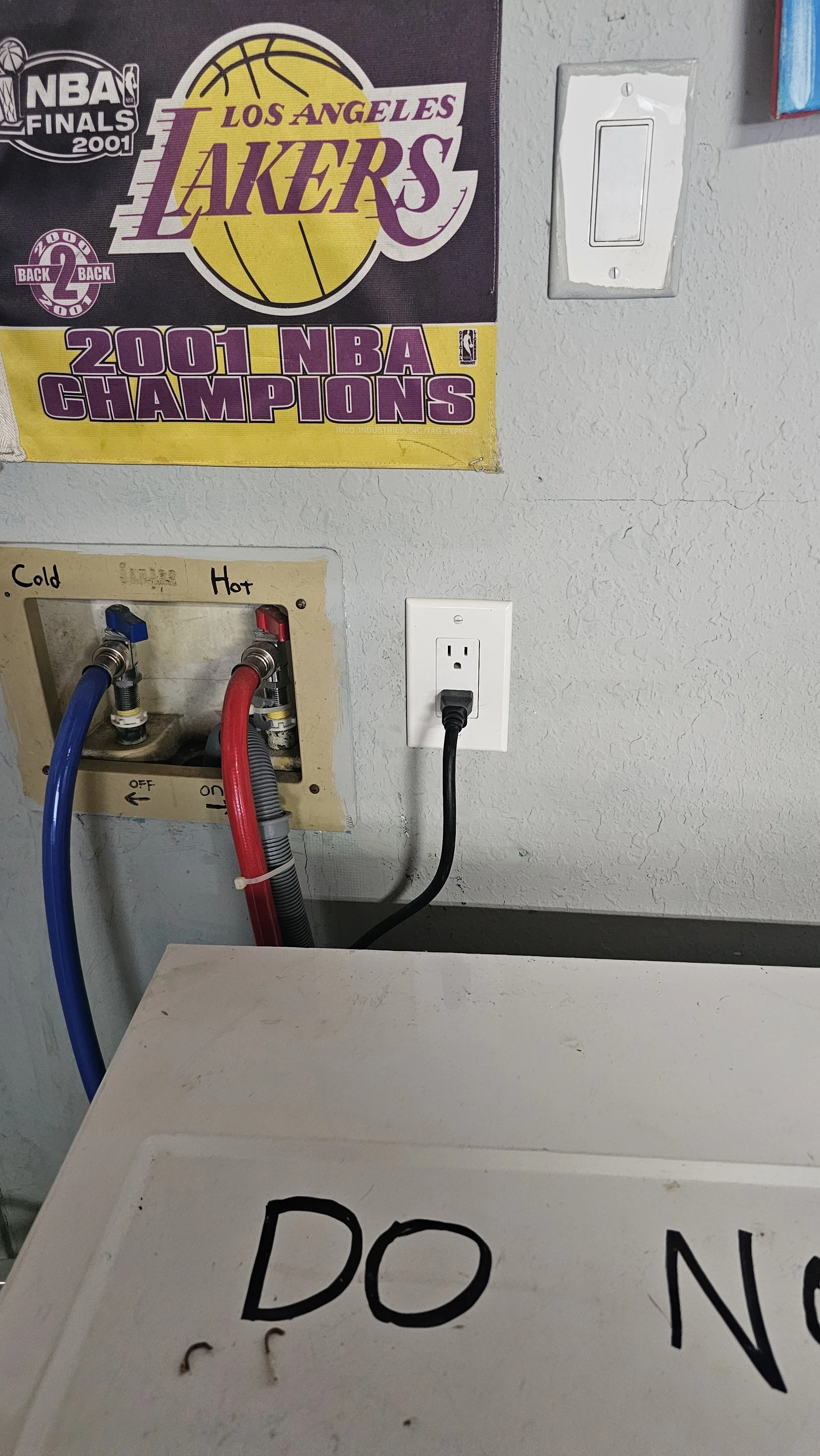 Wall with a Los Angeles Lakers poster highlighting their 2001 NBA championship, a standard electrical outlet with a black plugged-in cord, two water pipes labeled 'Cold' and 'Hot' connected to a box, and a partially visible white sign with black text that says 'DO N'.