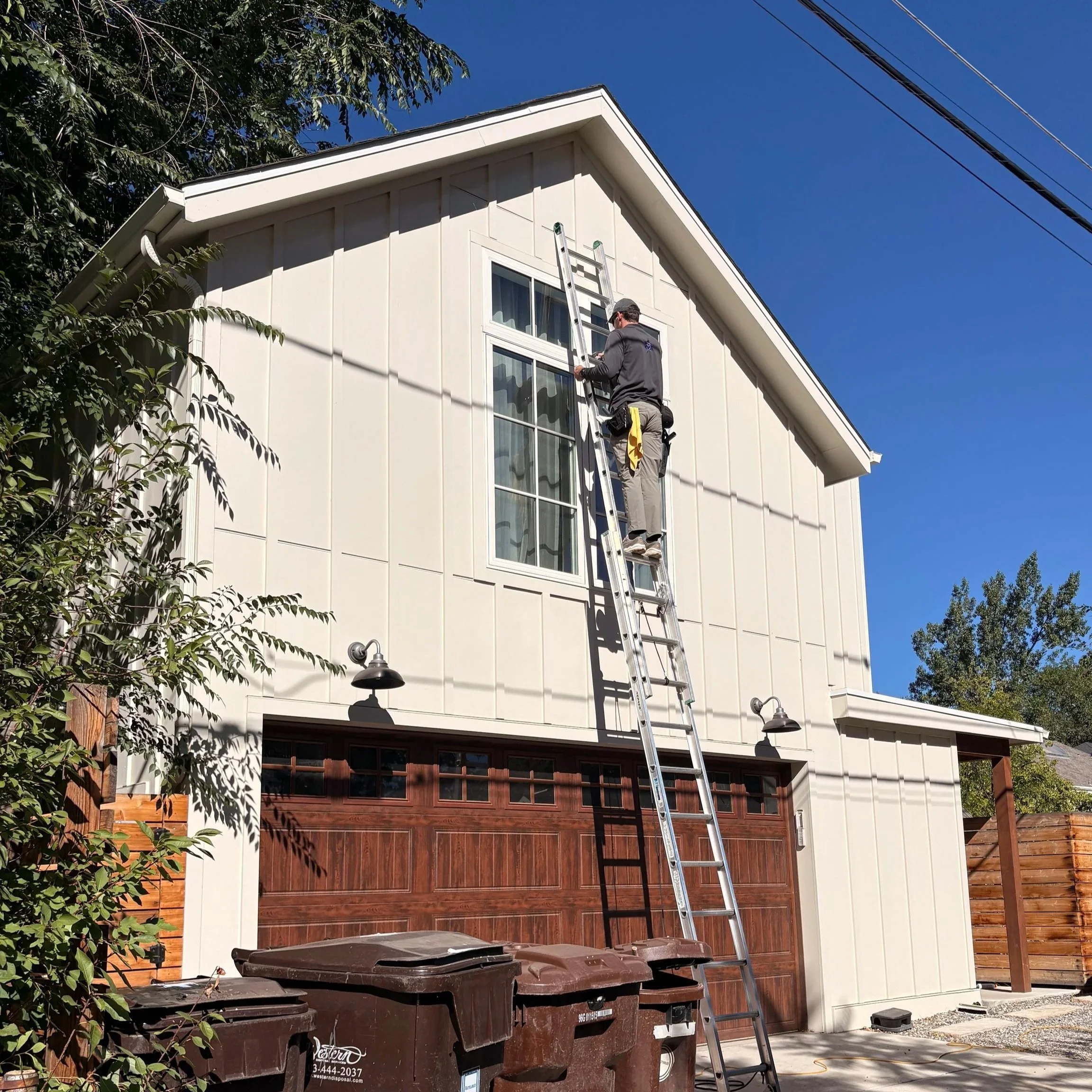 A technician working on the exterior windows of an accessory dwelling (ADU) in Boulder, standing on a ladder, under a clear blue sky, with trees and power lines in the background.