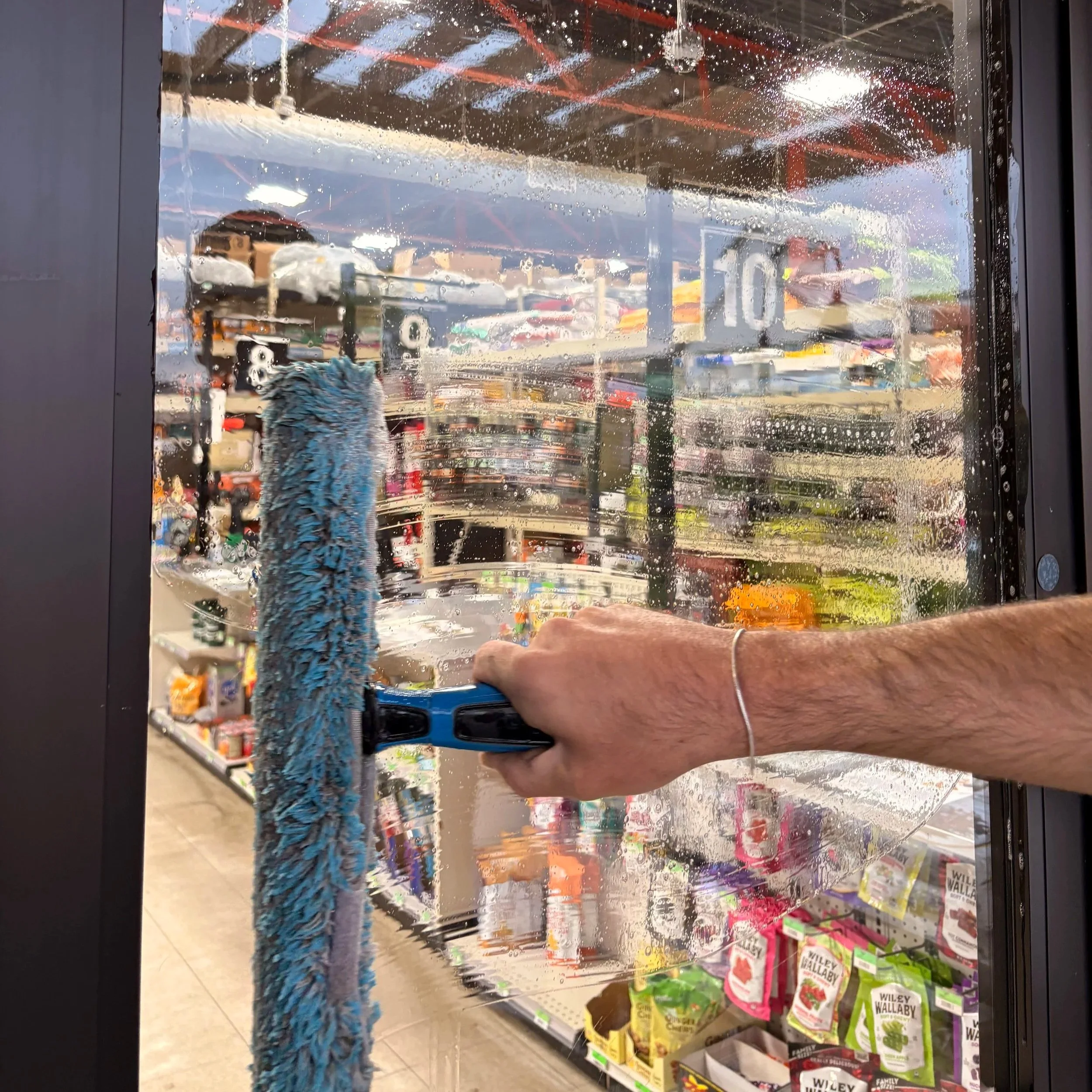 View through a glass window with water and soap droplets, showing a store aisle inside with shelves of gardening supplies and bags of plant soil. Technician hand holding a soapy squeegee. 