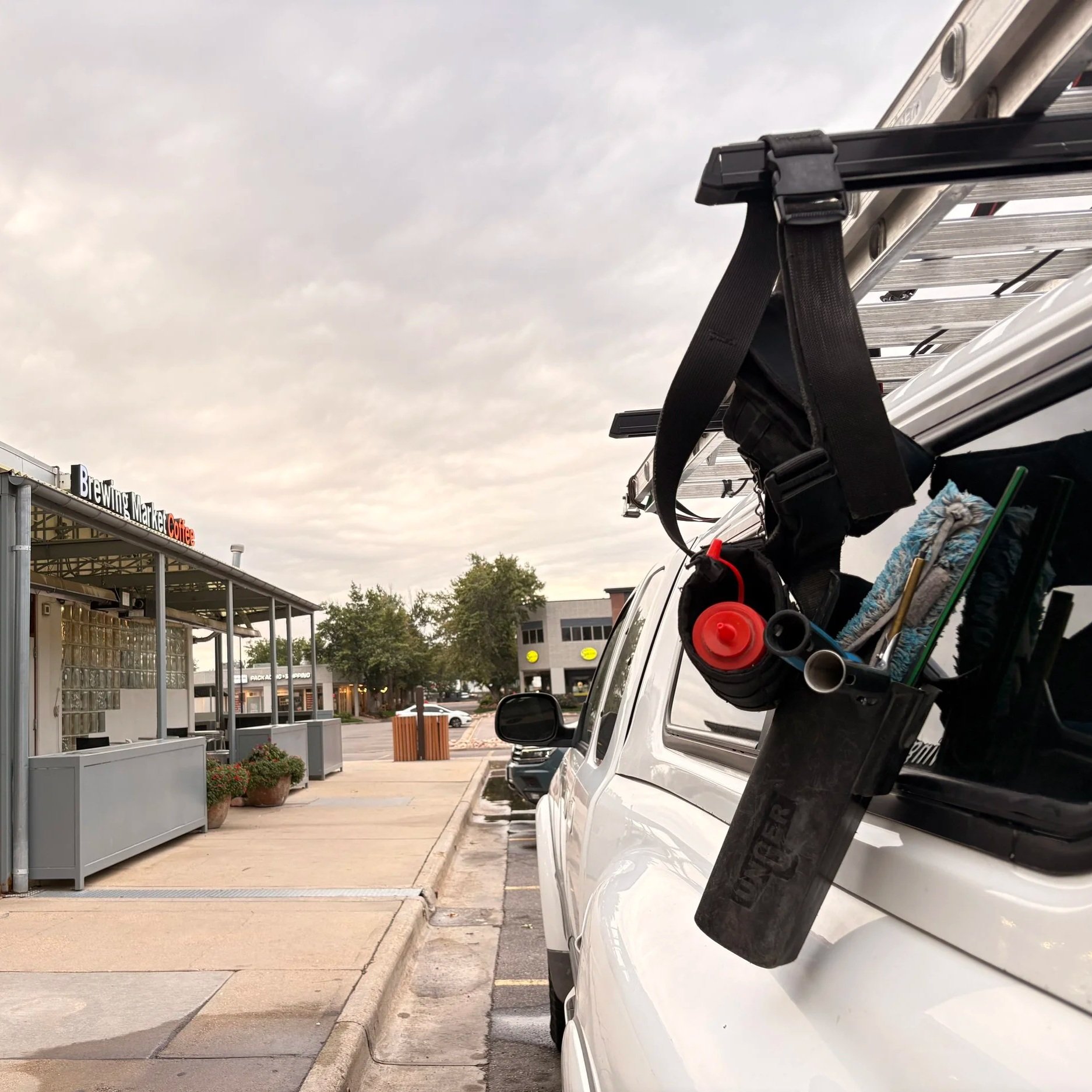 View of a parking lot outside a shopping center with multiple parked cars, a ladder extending from a truck, and a storefront sign reading 'Brewings Market Coffee' under a cloudy sky.