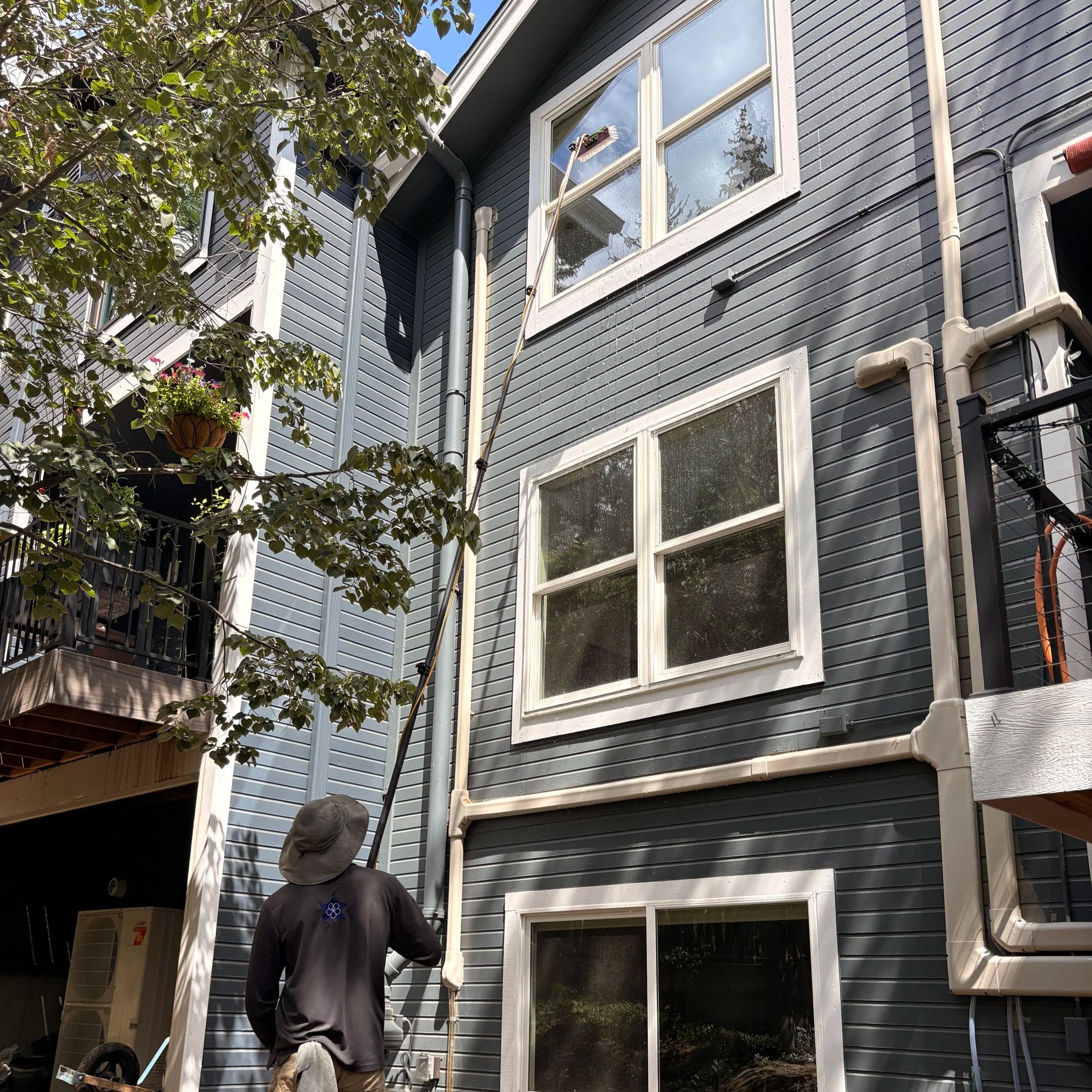A man cleaning the exterior window of a blue apartment with a water-fed pole while standing outside. The apartment has multiple windows and gray-blue vertical siding. 
