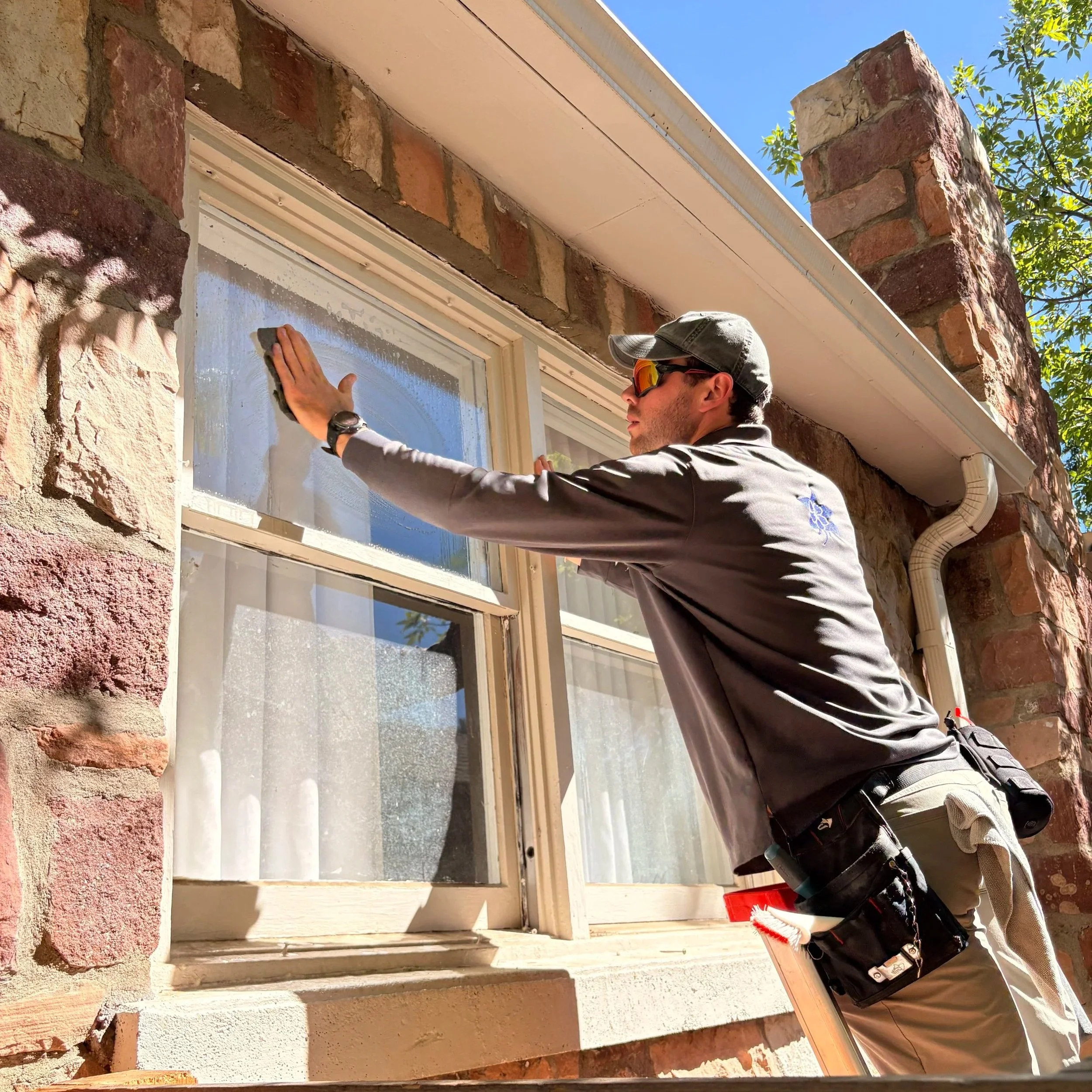 A technician wearing sunglasses, a gray cap, and uniform is cleaning storm windows of a historic Boulder bungalow home while standing in a ladder. The window is set in a brick wall, and there is blue sky and green foliage in the background.