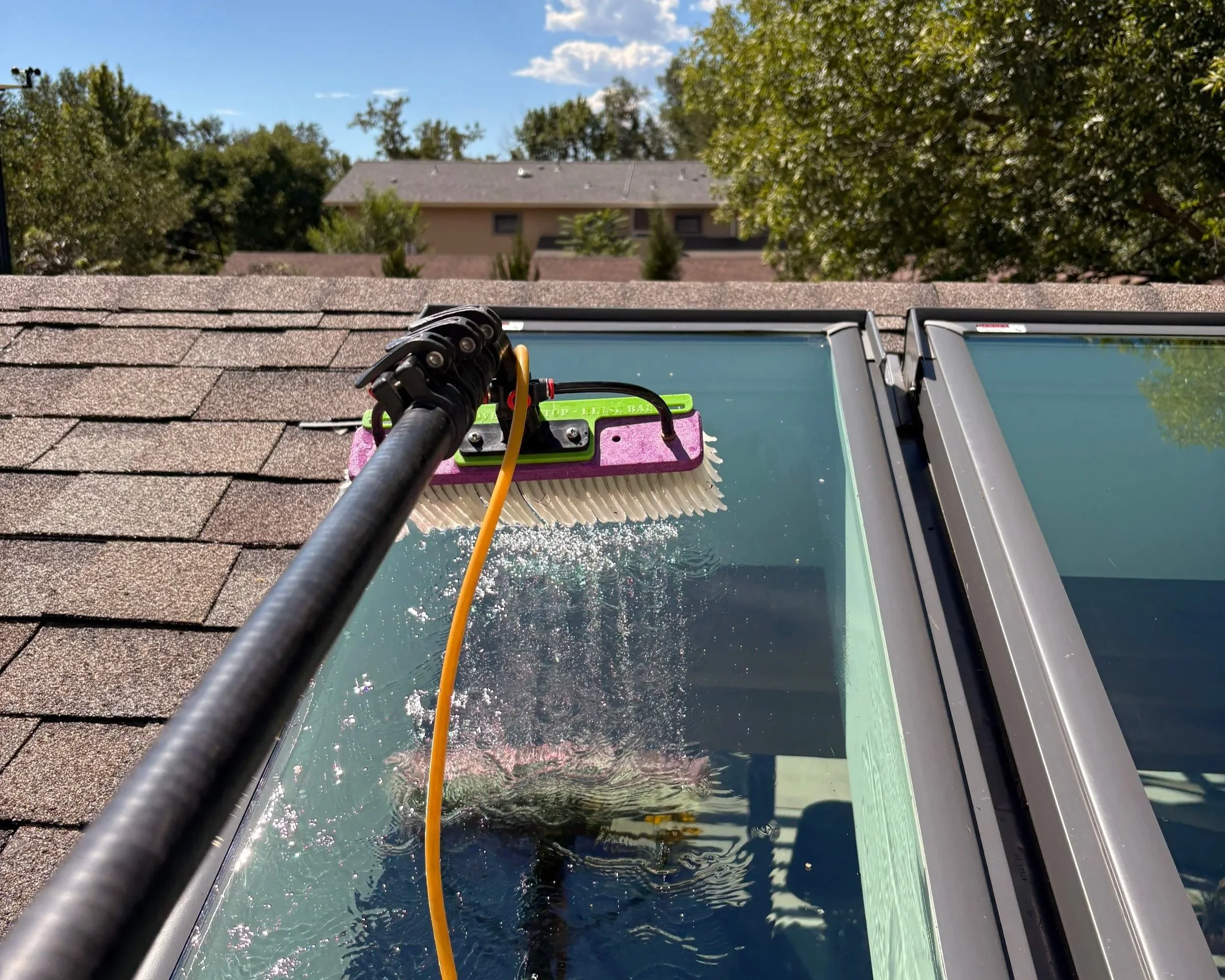View of a roof with sky lights, showing cleaning equipment including a scrub brush, hose, and water-fed pole with a background of trees, houses, blue sky, and clouds in Erie, Colorado.