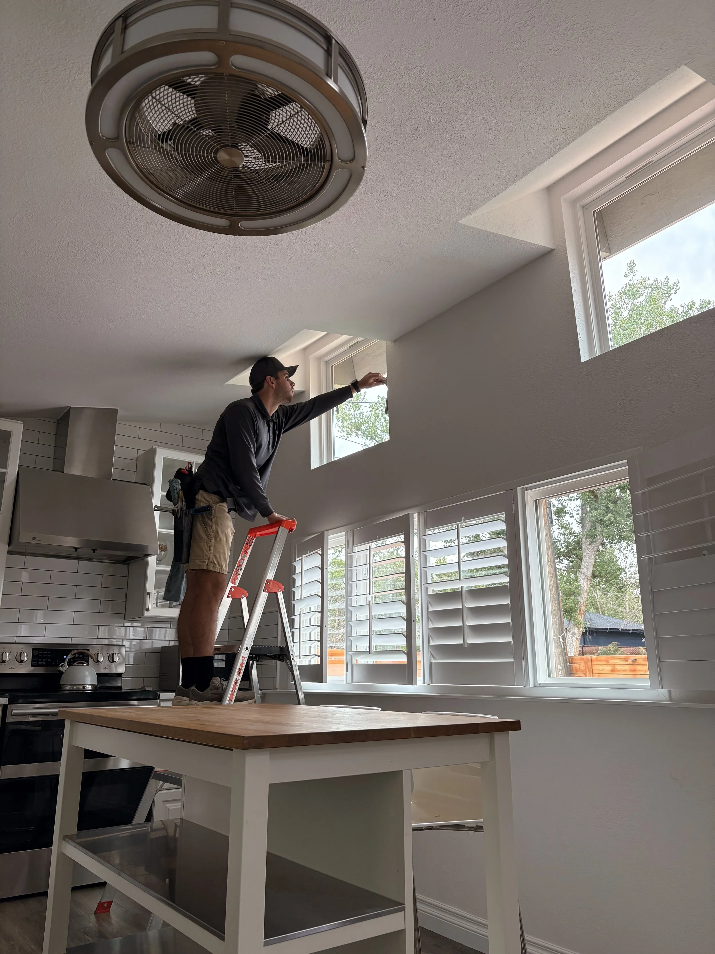 A man standing on a step ladder cleaning windows in a bright kitchen with white cabinetry and a window showing trees outside in Superior, Colorado.