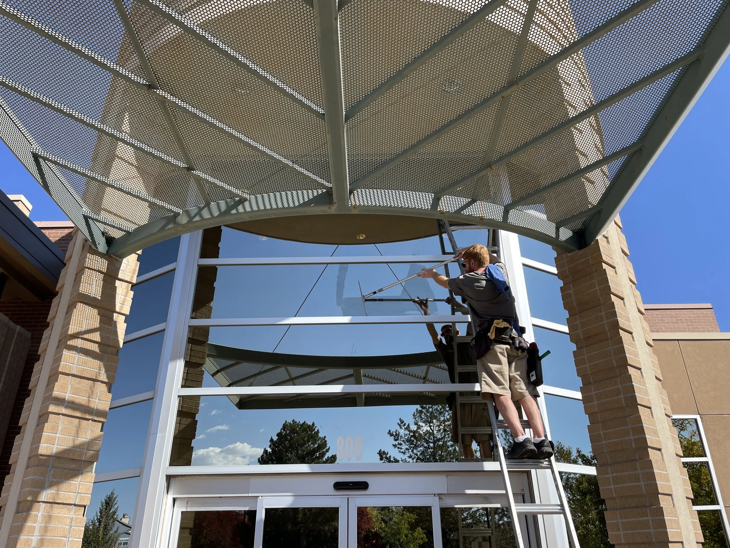 Technician standing on a ladder cleaning the exterior window frame beneath a large metal awning on a brick building.