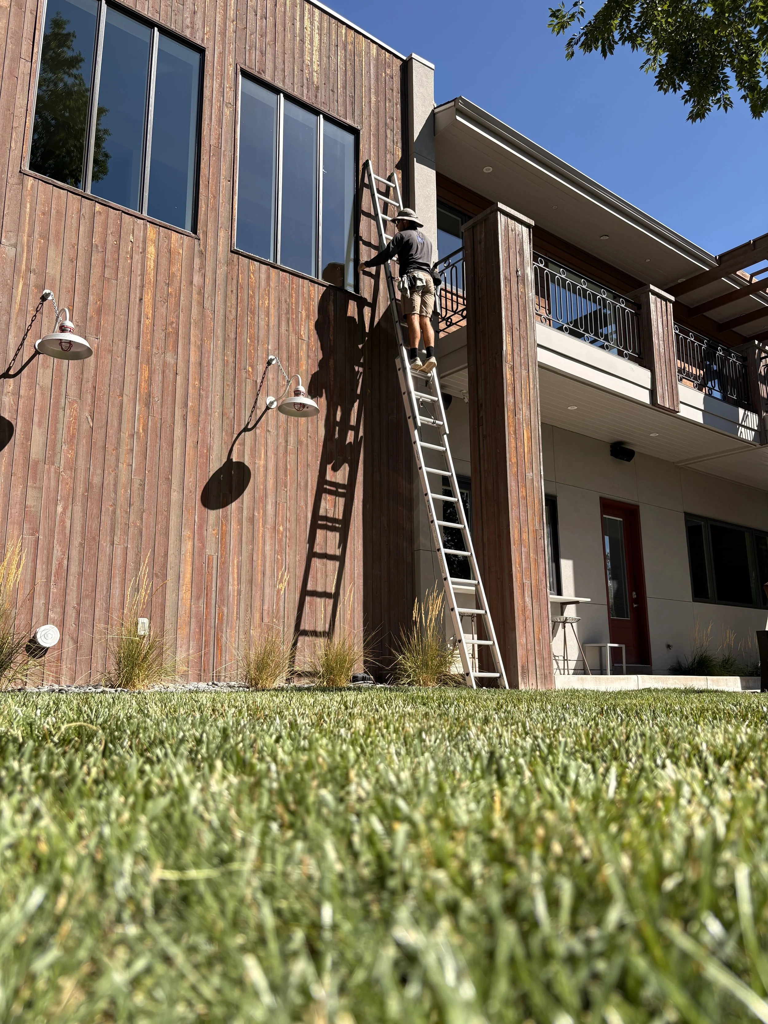 A technician standing on a tall ladder working on the exterior of a modern building with large windows, wooden siding, and a balcony, under a clear blue sky.