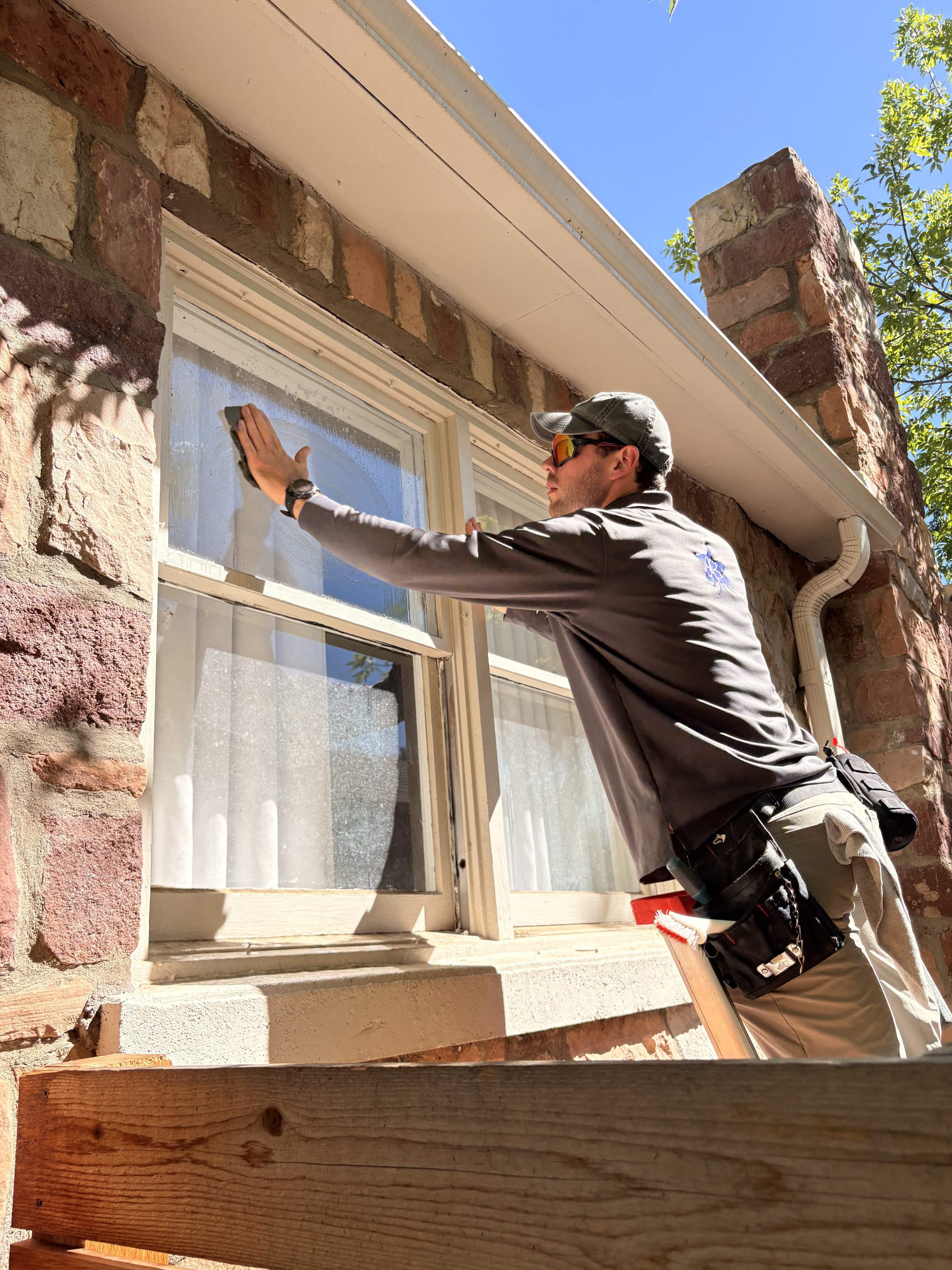 A technician wearing sunglasses, a gray cap, and uniform is cleaning storm windows of a historic Boulder bungalow home while standing in a ladder. The window is set in a brick wall, and there is blue sky and green foliage in the background.
