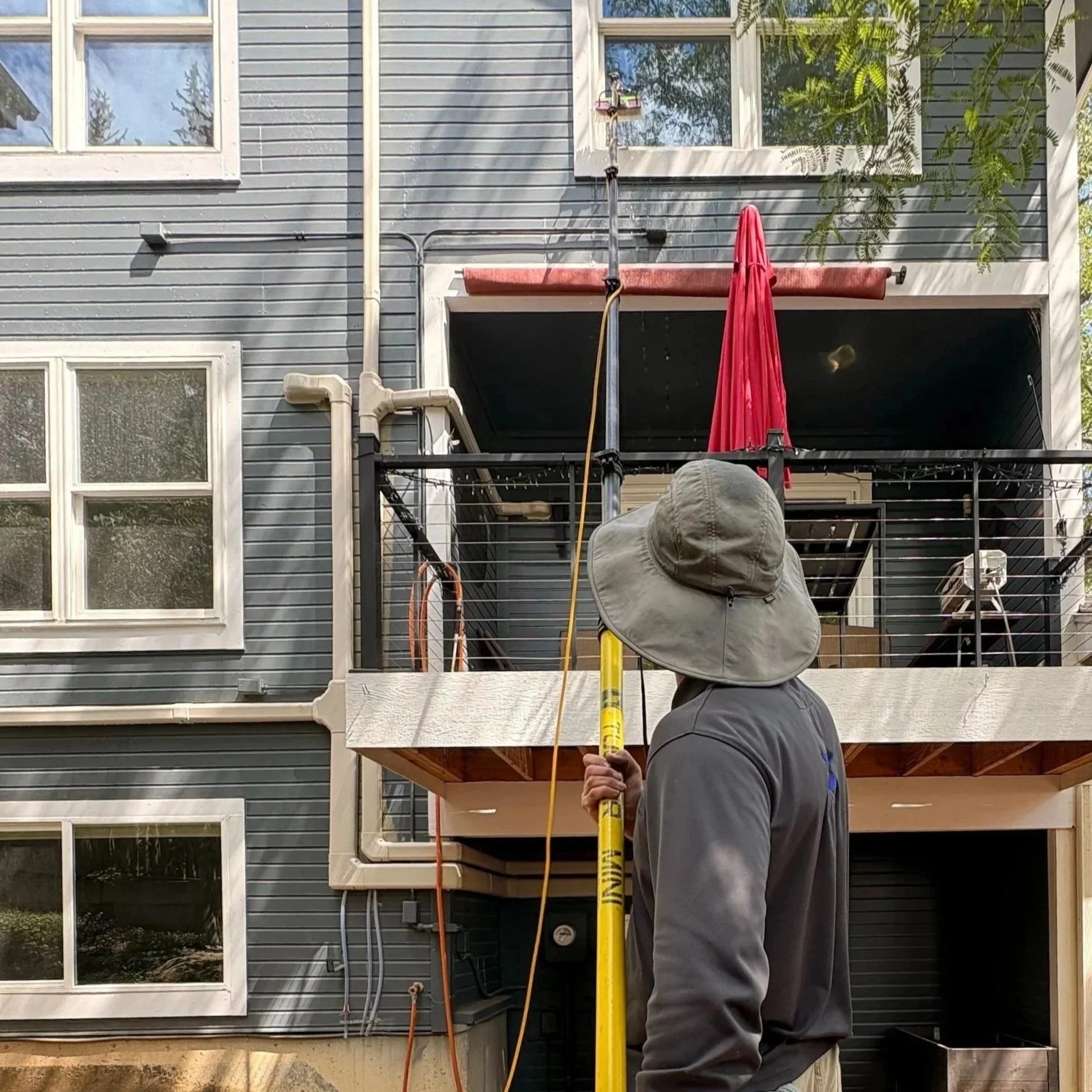 Worker in uniform and sun hat holding a yellow water-fed pole, in front of a blue apartment with multiple windows and trees in the background.