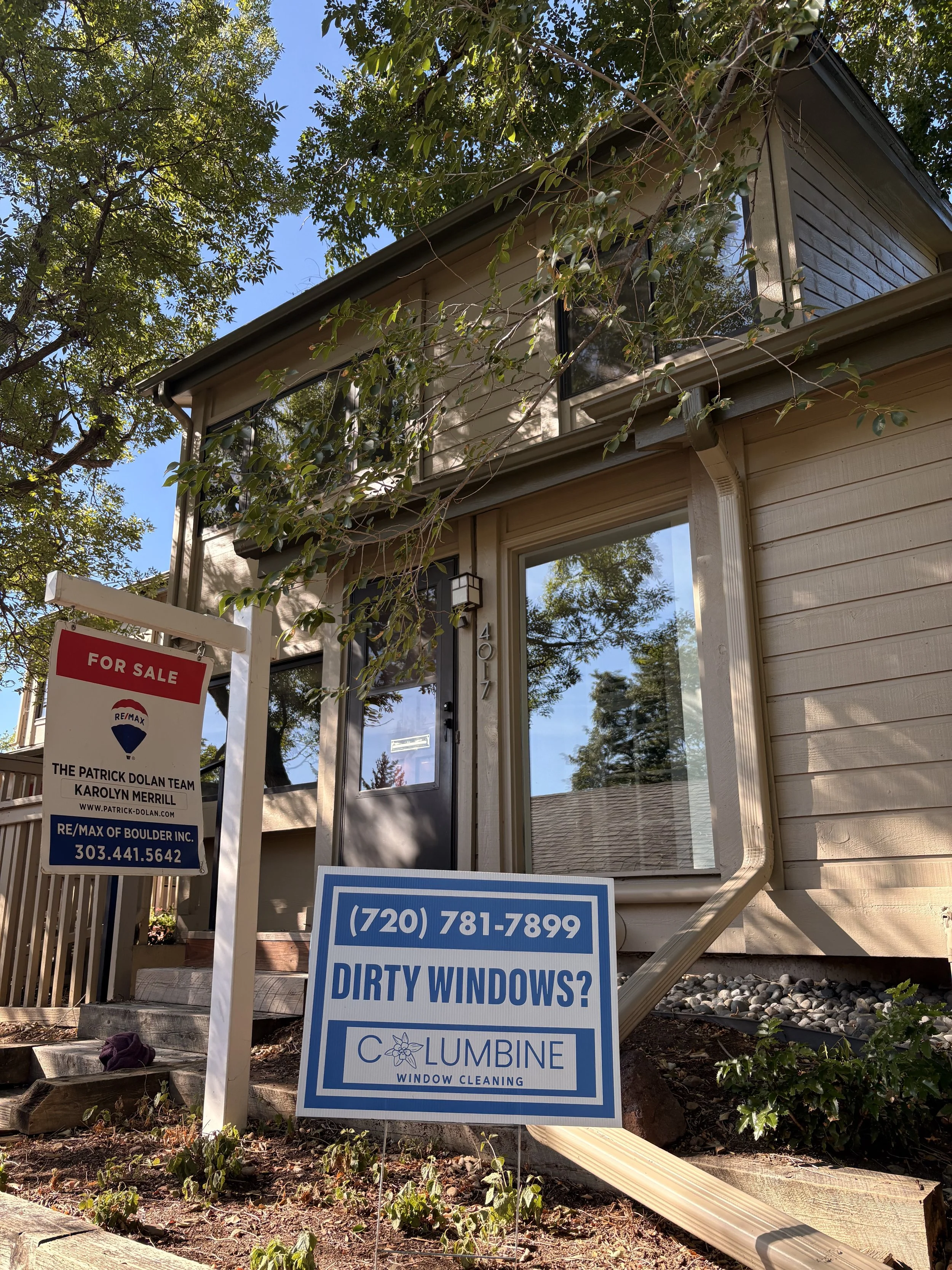 House with a for sale and window cleaning service signs in front, trees and a clear sky in the background.