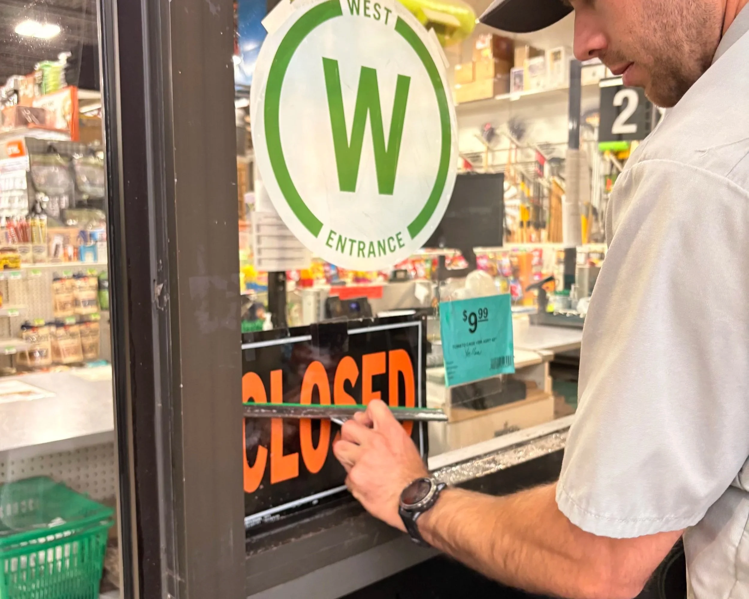 A  technician is cleaning the glass door of a Boulder hardware store with a professional squeegee. Inside the store, shelves stocked with various products can be seen.
