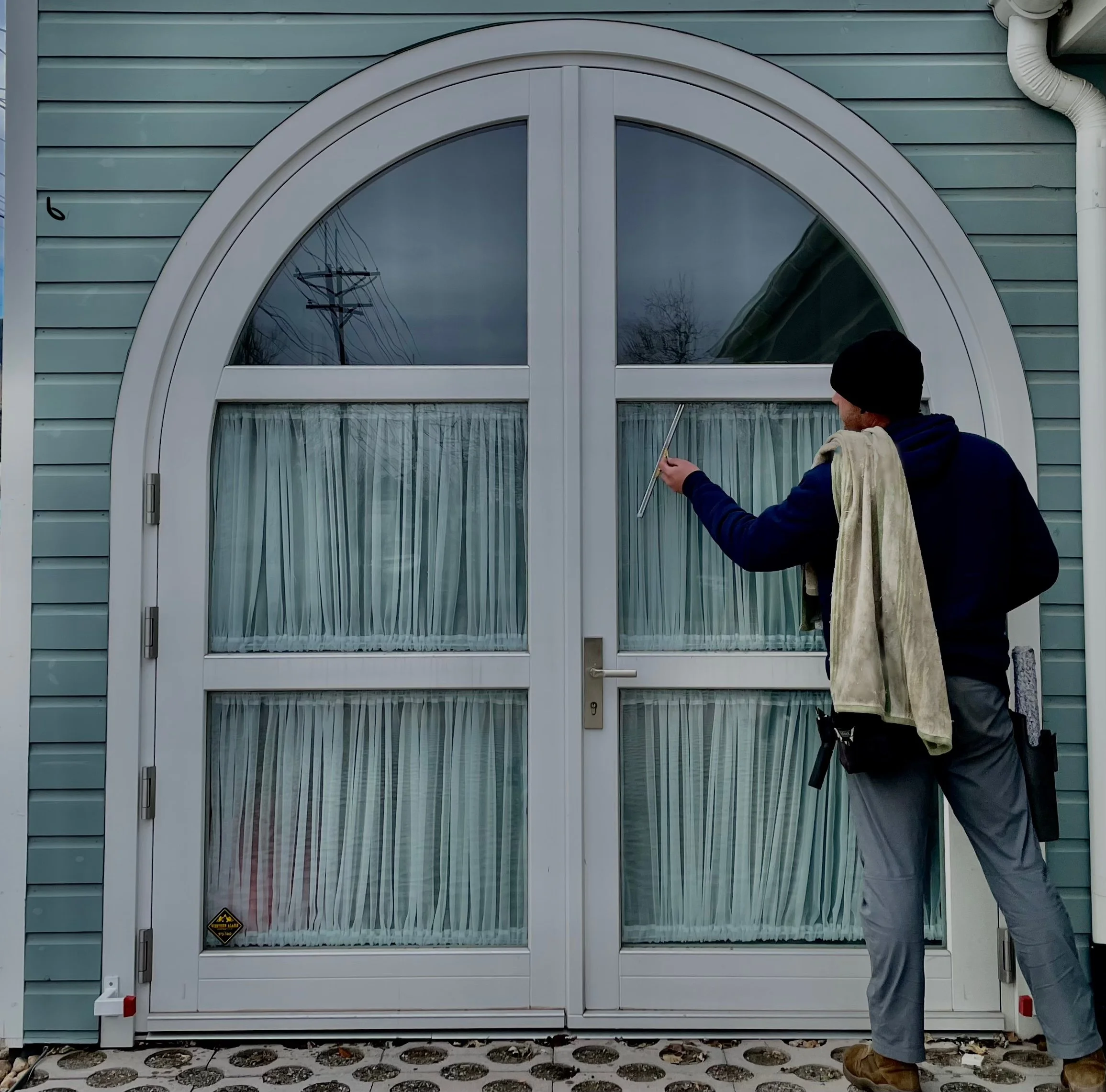 Worker cleaning a large glass door with a cloth and squeegee outside a house with blue siding.