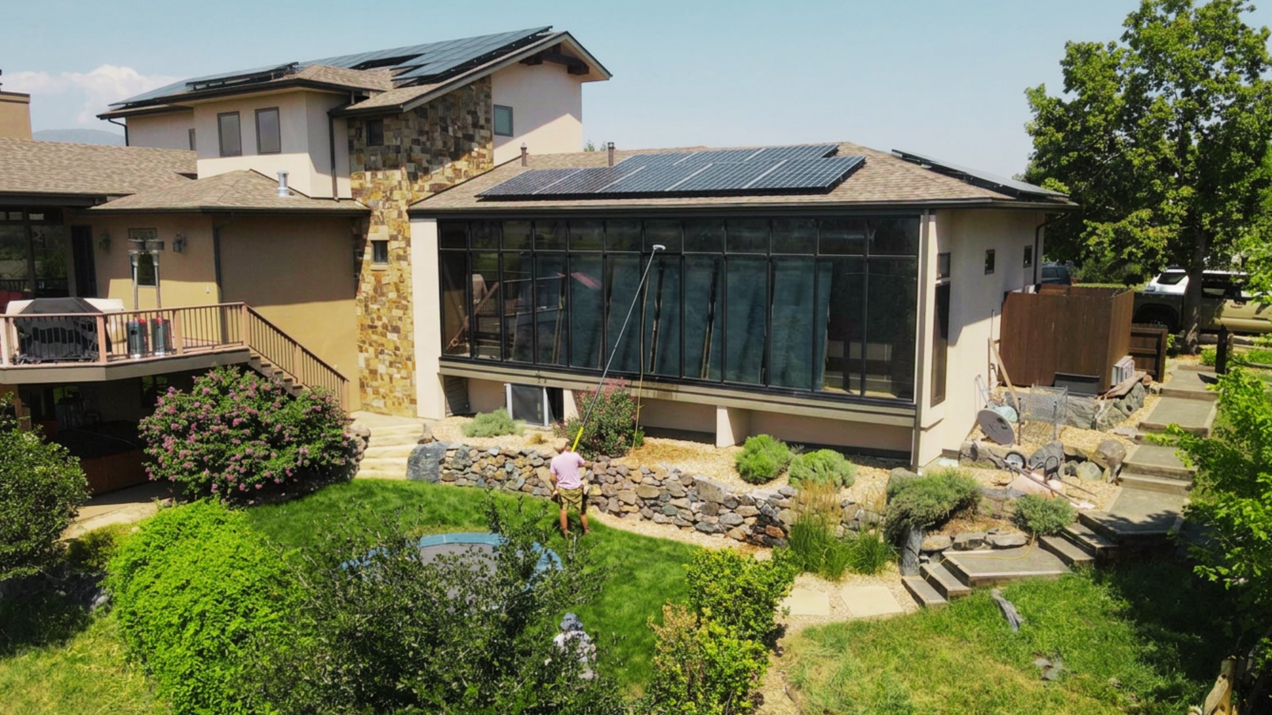 A house with solar panels on the roof and a glass-enclosed porch on the back. The yard has green grass, bushes, and trees, with a technician working on large panoramic floor-to-ceiling windows in the garden.