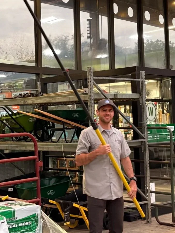 A technician standing outside a garden center, holding an extended water-fed pole, smiling at the camera. The background shows shelves with gardening supplies like wheelbarrows and bags of soil.
