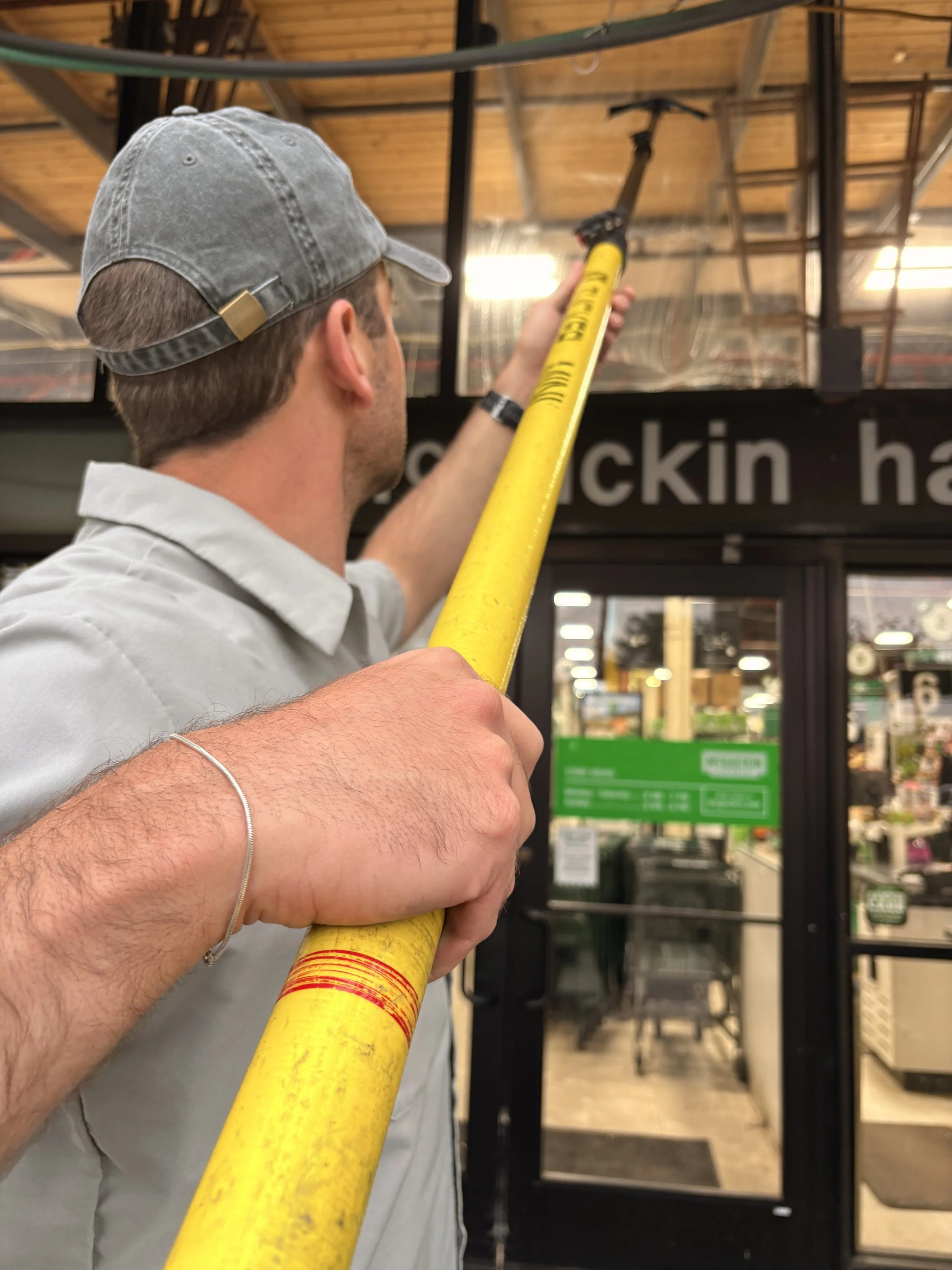 A man wearing a gray cap and gray uniform is cleaning a retail shop exterior with a yellow extension water-fed pole.
