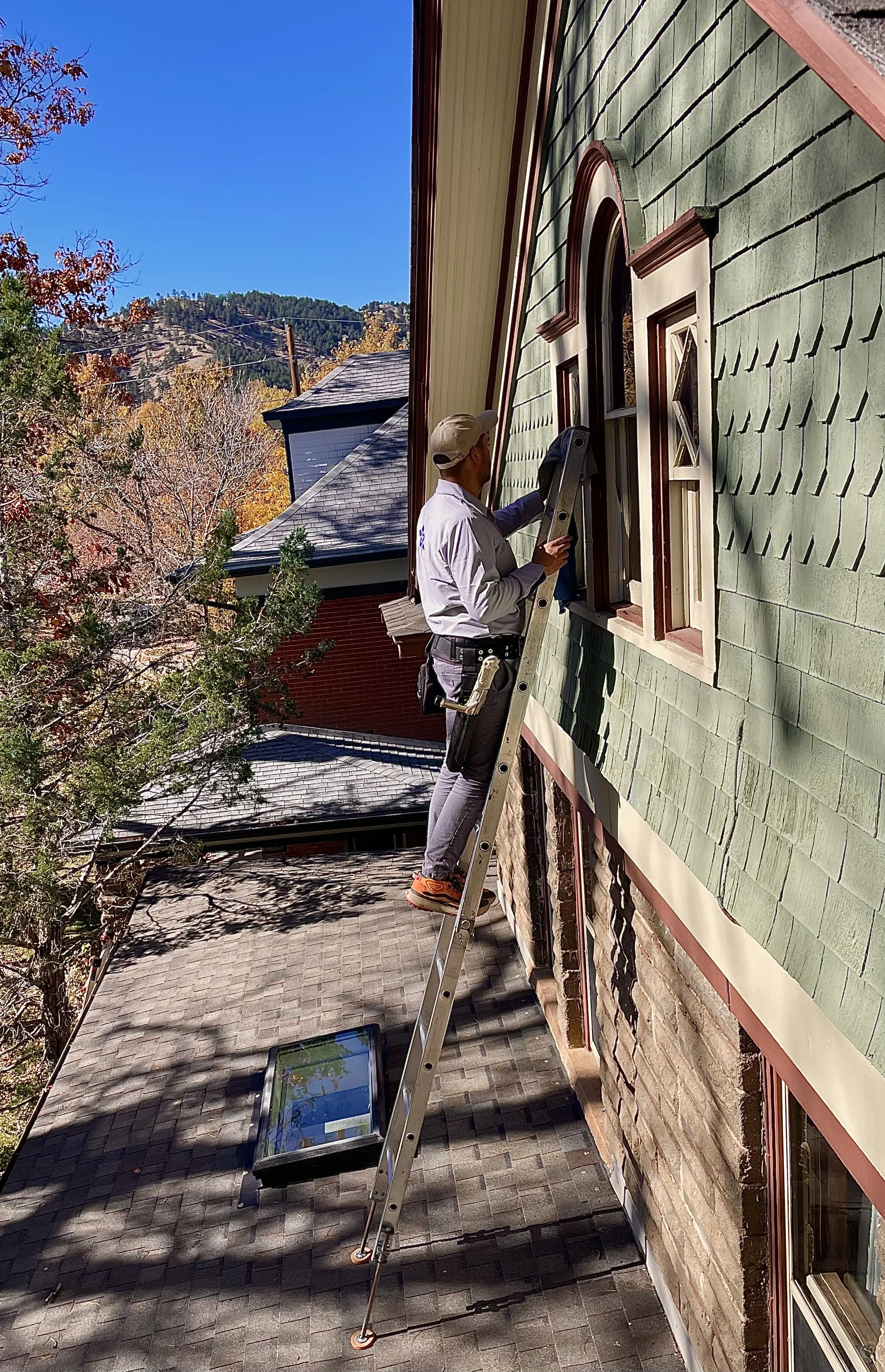 A man on a ladder cleaning a window on the second story of a green house. The scene is outdoors with trees and hills in the background under a clear blue sky.