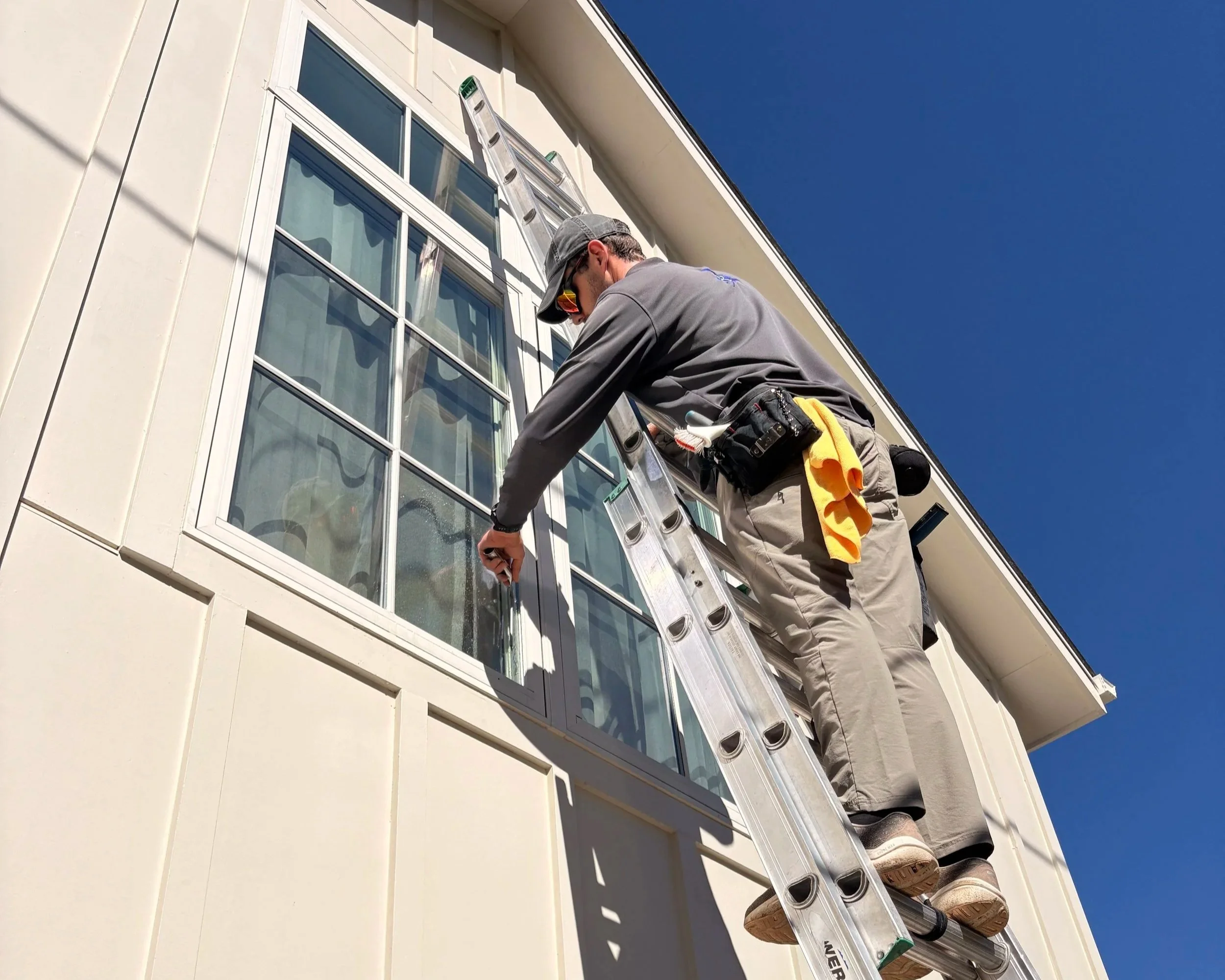 A technician wearing a uniform is standing on a ladder, cleaning on the large window of a modern white building with multiple panes. The sky is clear and blue.