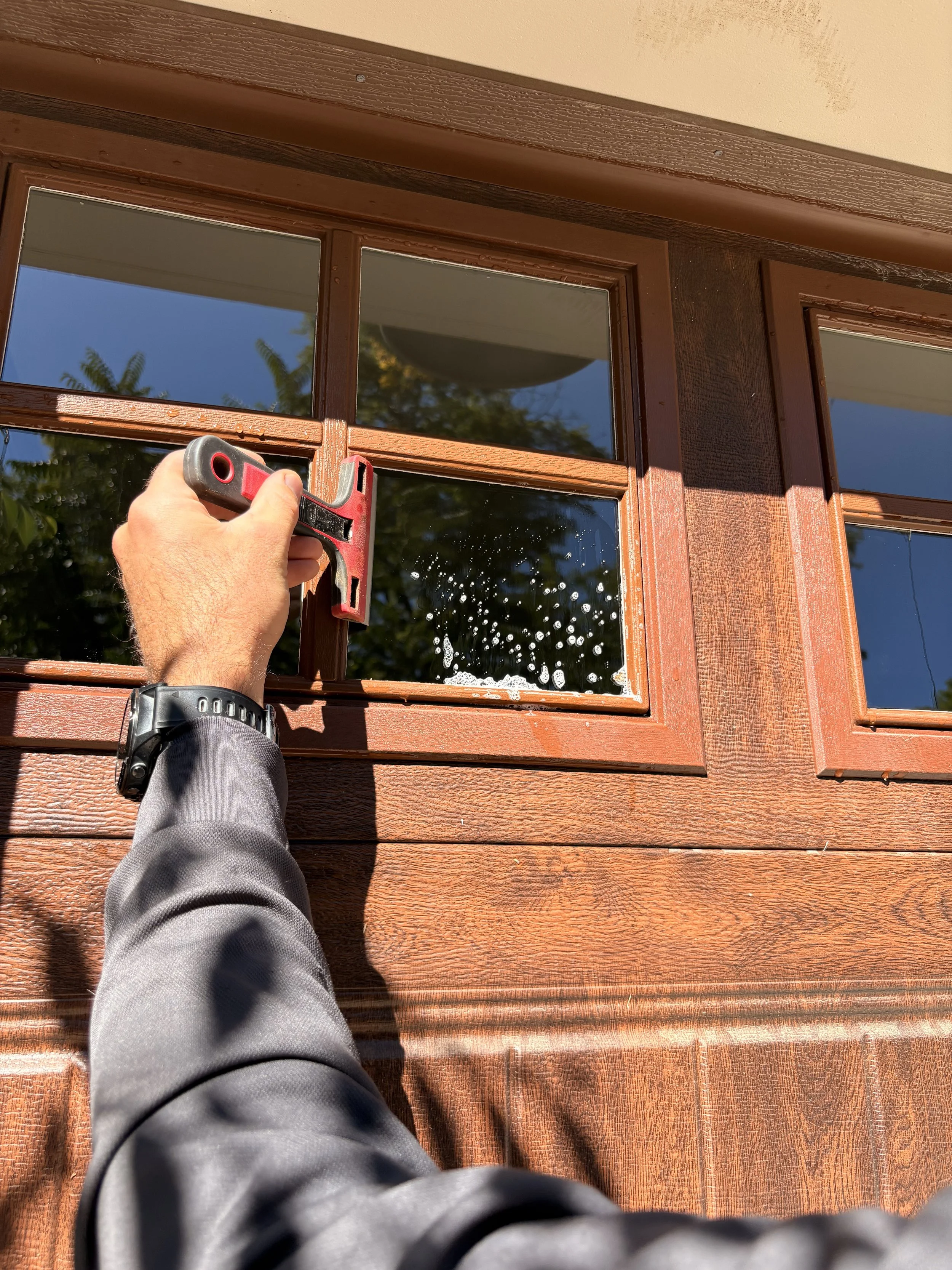 A hand is cleaning a garage door window with a squeegee under sunlight.