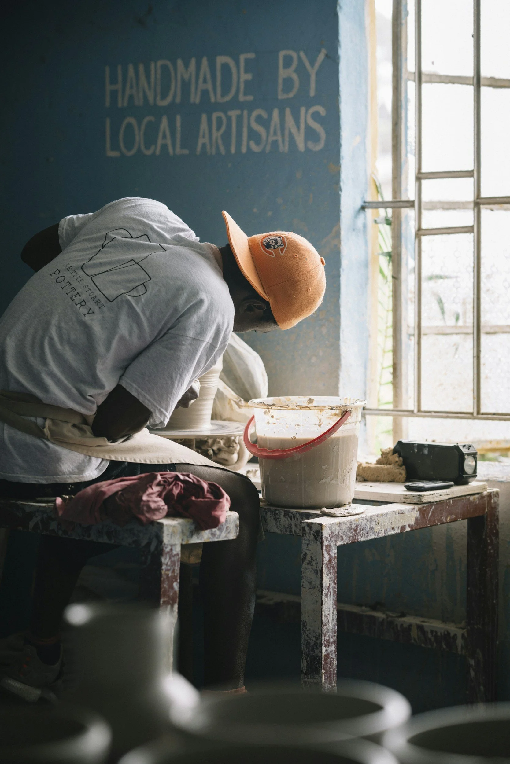 Person working on pottery in a workshop with a blue wall that reads 'Handmade by Local Artisans,' sunlight coming through a window.