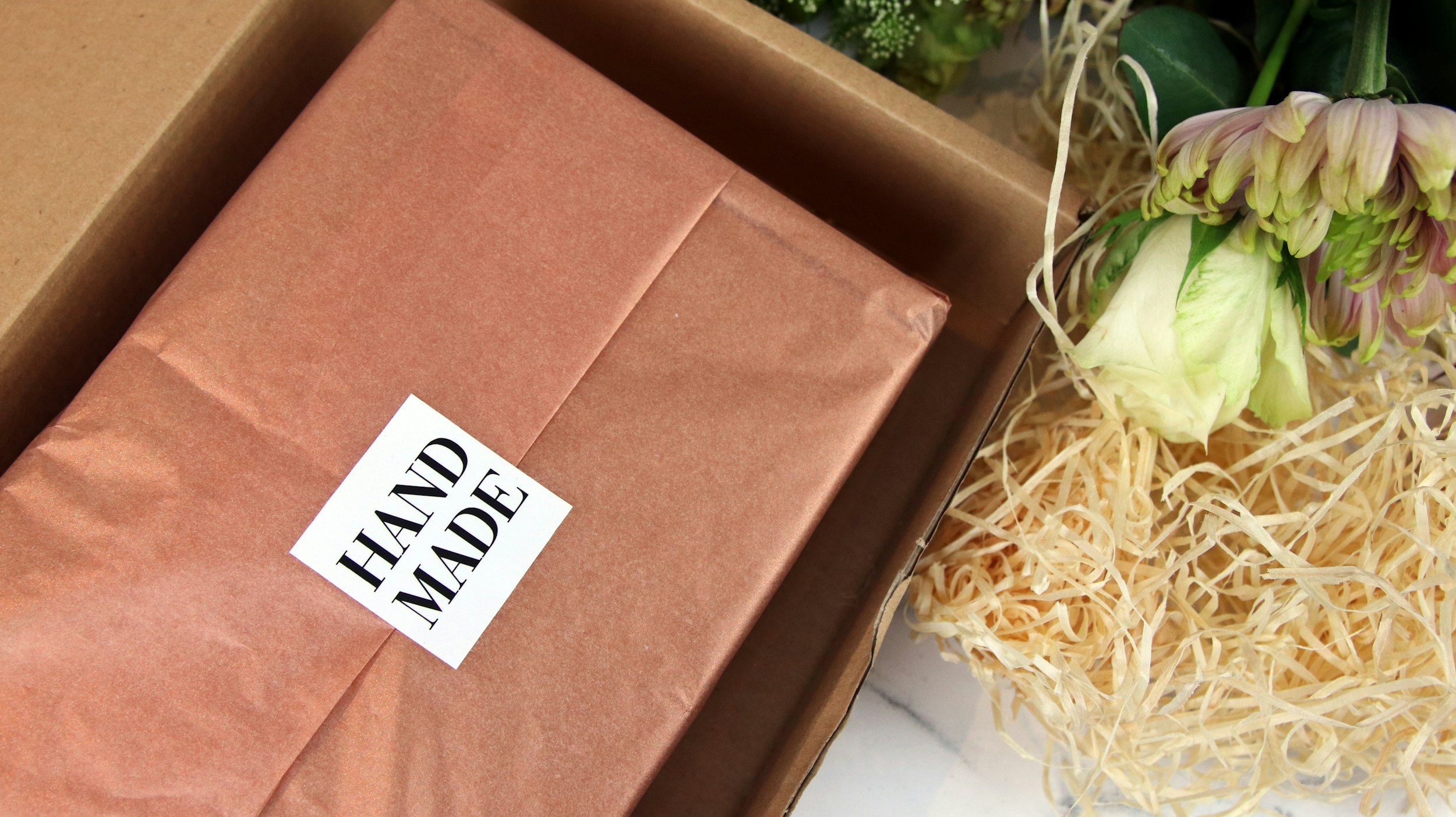 Close-up of a gift wrapped in brown paper with a "Handmade" sticker on it, surrounded by crinkled paper shred and a bunch of green and pink flowers.