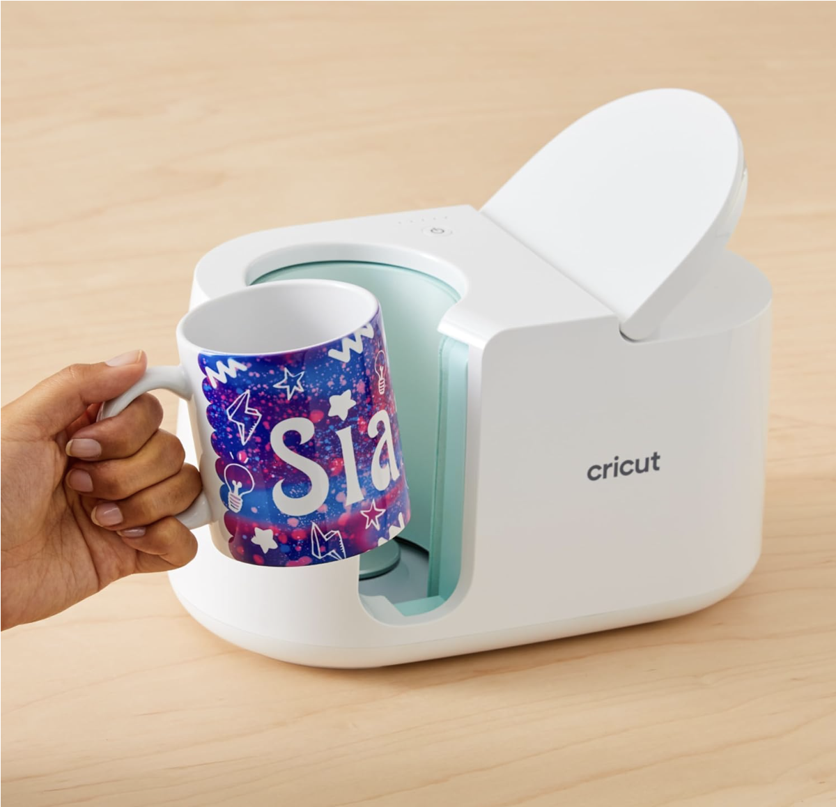 A person placing a mug with a colorful design and the word 'sia' into a white Cricut mug press machine.