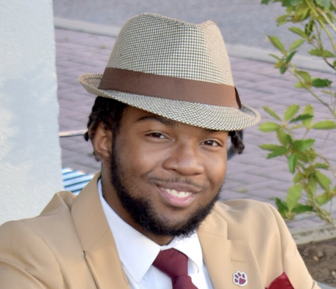 A young man with a beard smiling while wearing a beige blazer, white shirt, maroon tie, and a tan fedora hat with a brown band. He is sitting outdoors next to a white wall and some green plants.