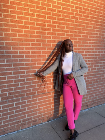 Woman with long braids leaning against a brick wall, wearing a gray blazer, white shirt, bright pink pants, and black shoes.