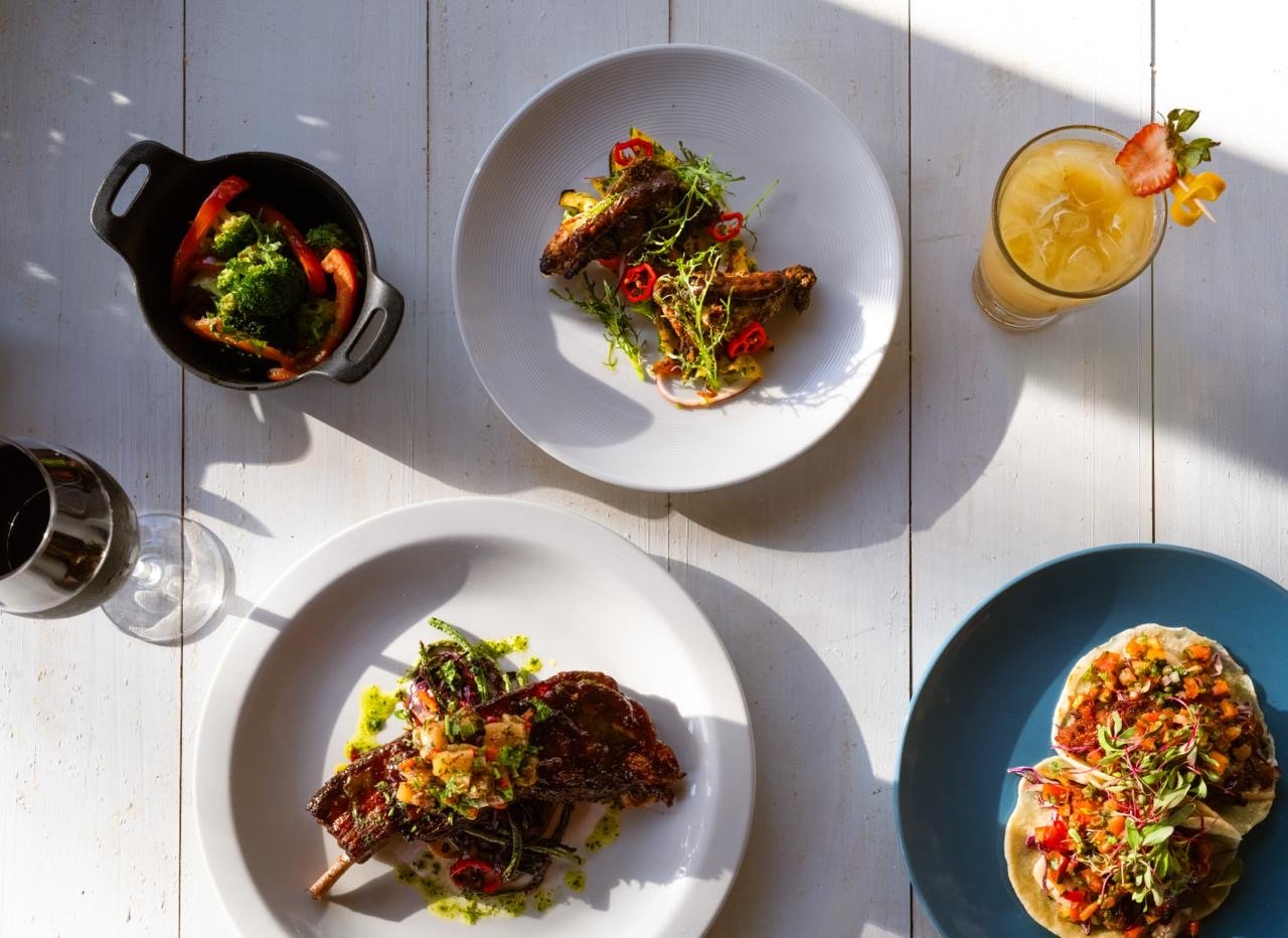 Overhead view of a white wooden table with various dishes, including a plate of grilled ribs with sauce and herbs, a roasted chicken leg with vegetables, a tostada with toppings, a bowl of vegetable stir-fry, and a glass of pineapple cocktail with a strawberry garnish.