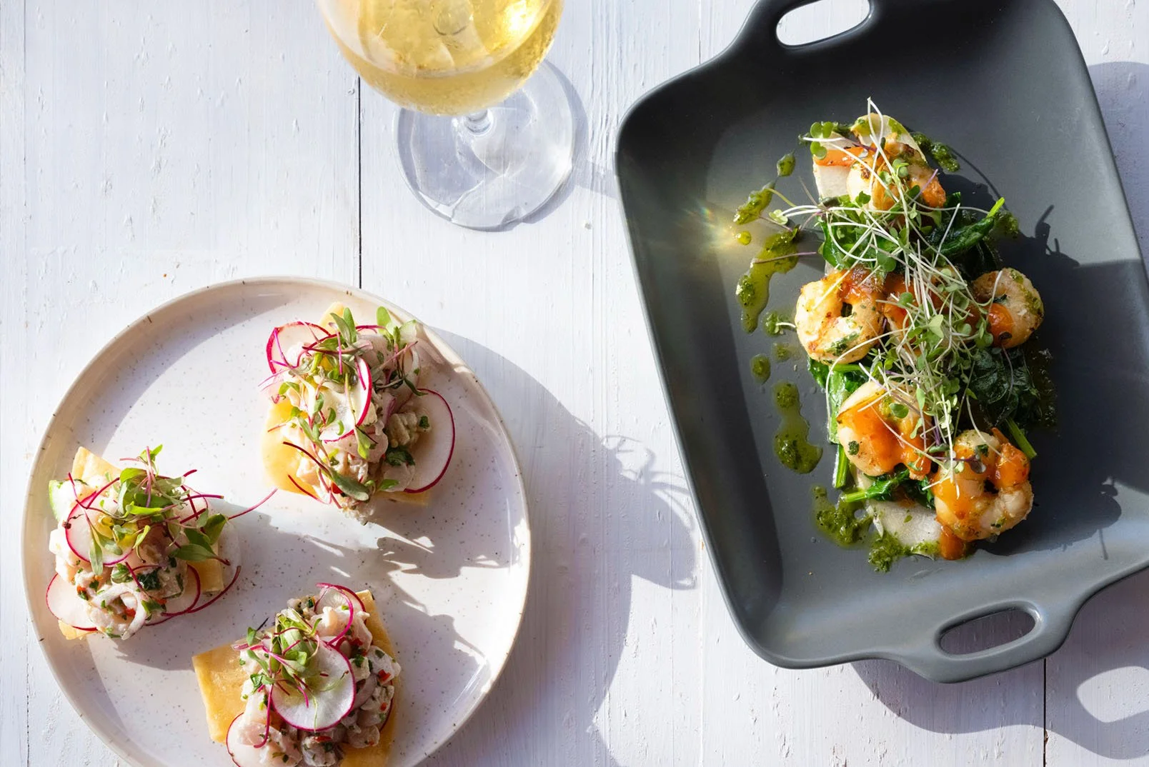 Plate of small open-faced sandwiches topped with radish slices, microgreens, and cream, alongside a black serving dish with a seafood and vegetable dish garnished with microgreens, green drizzle, and lemon slices, and a glass of white wine on a white wooden table.