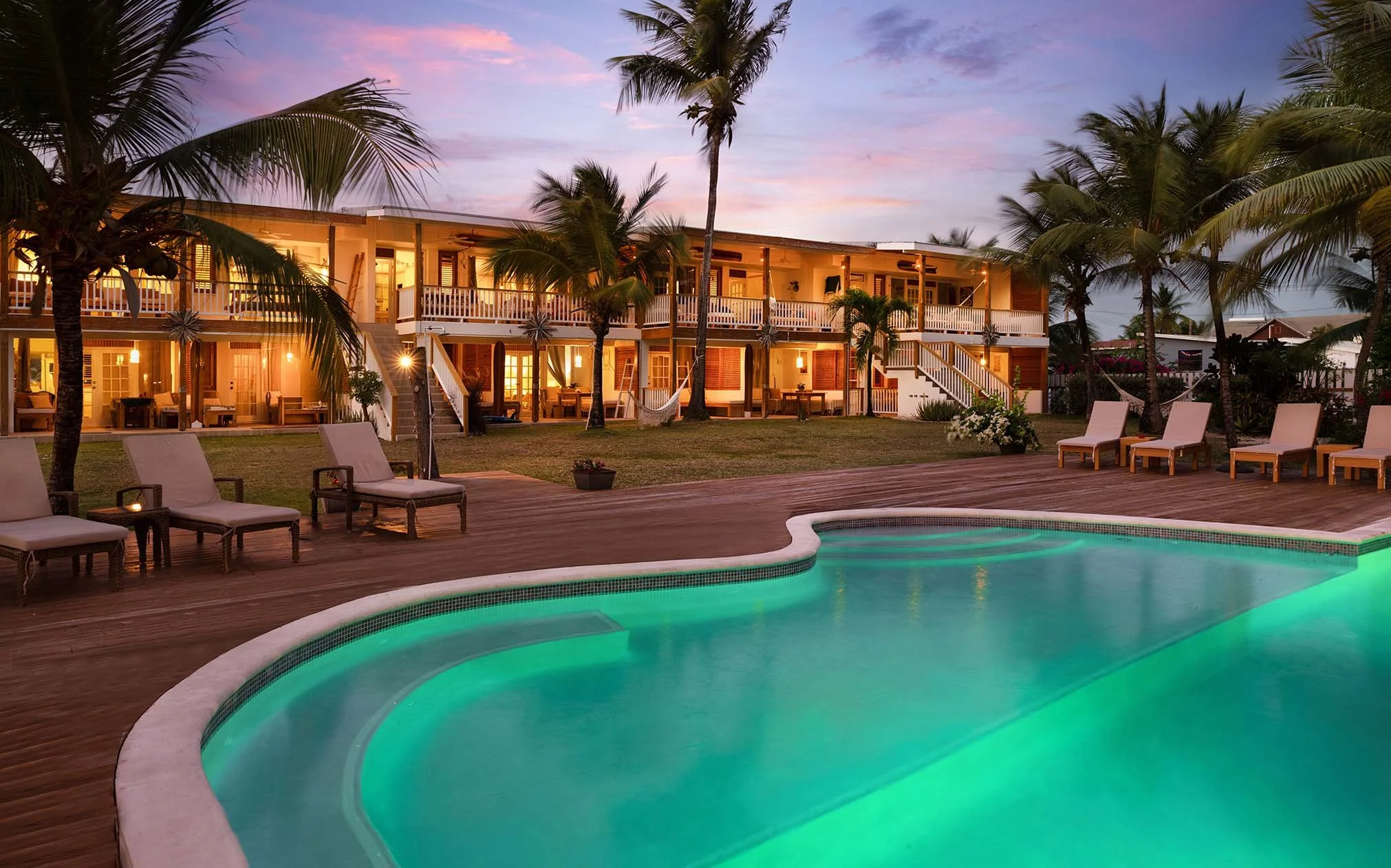 A luxurious tropical resort with a swimming pool illuminated by underwater lights, surrounded by wooden decking, lounge chairs, palm trees, and a two-story building with balconies and warm lighting, during sunset with a colorful sky.
