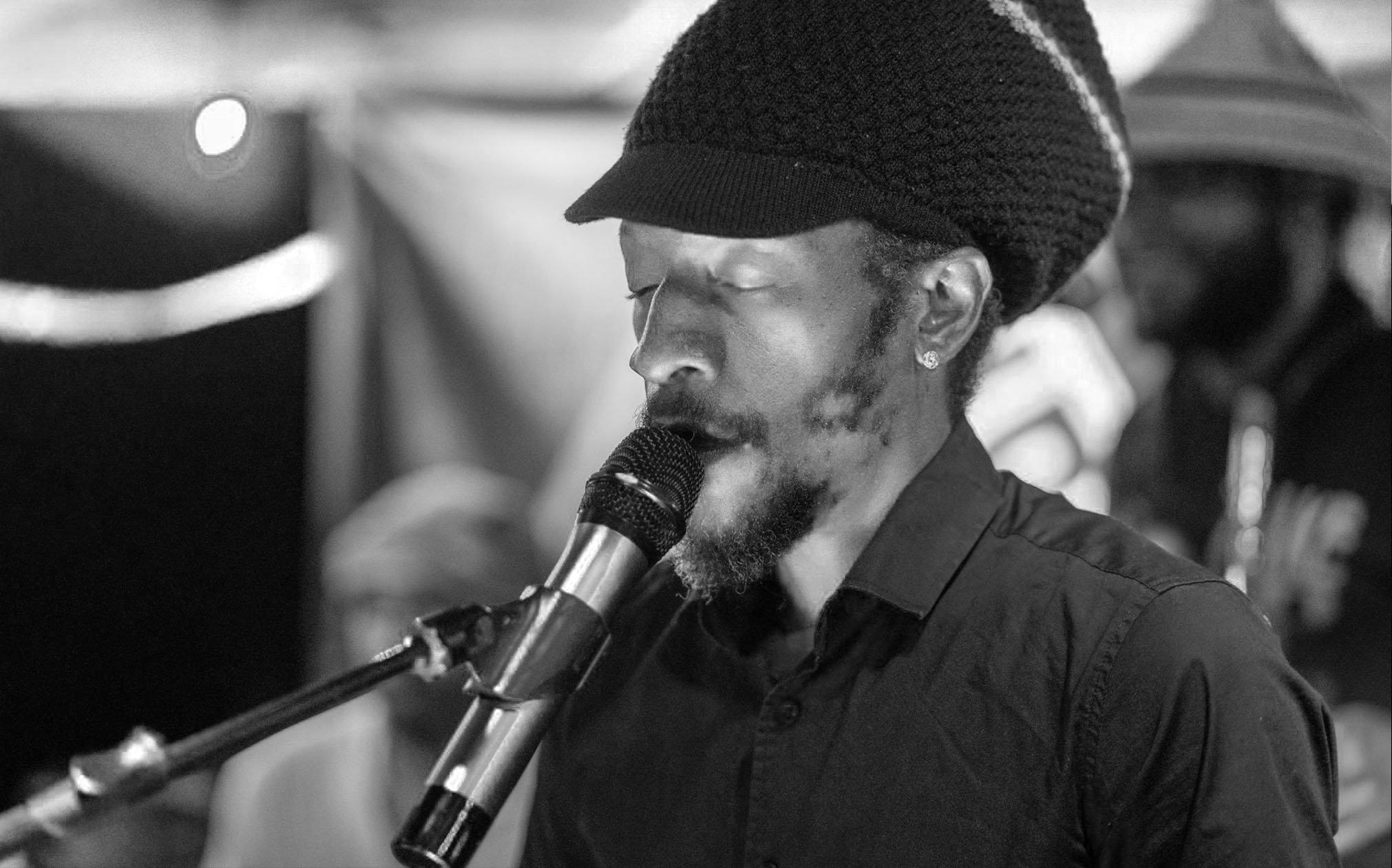 A man with a beard wearing a beanie and earrings singing into a microphone.