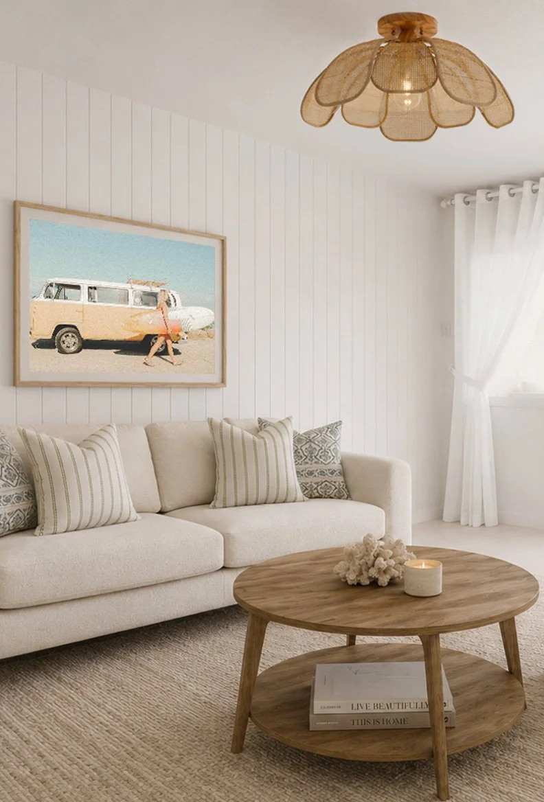 A cozy living room with a beige sofa decorated with striped and patterned pillows, a wooden coffee table with a candle and a decorative item on it, a framed beach-themed artwork on the white paneled wall, white curtains, and a ceiling light fixture resembling flower petals.