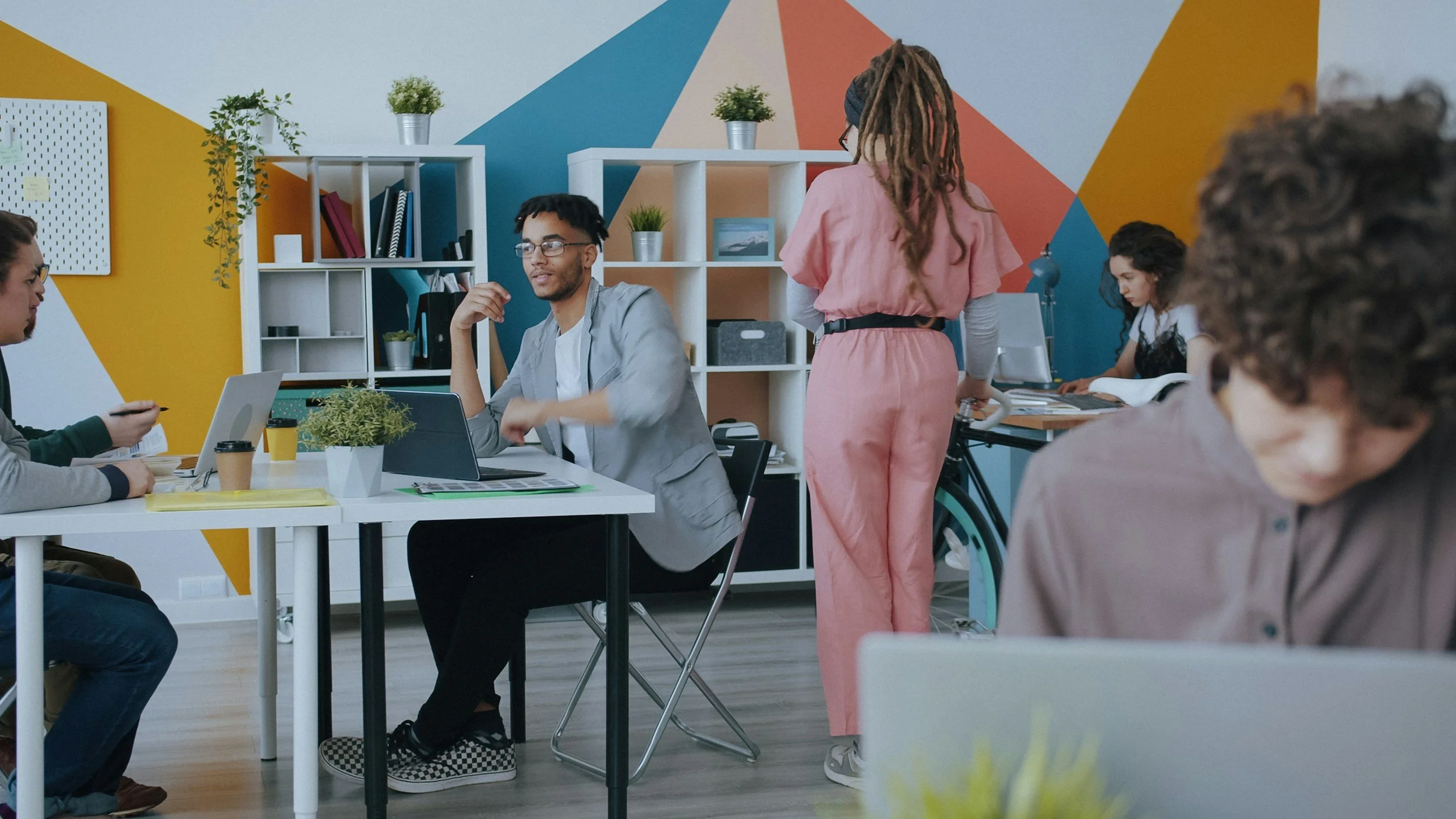 A diverse group of young people working together in a colorful, modern coworking space with shelves, plants, and a vibrant geometric wall mural.