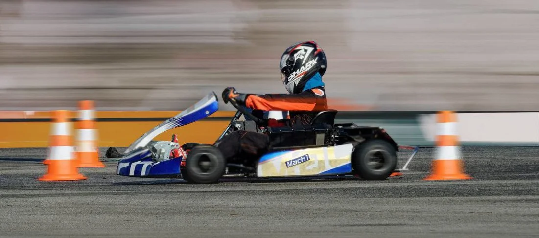 Ein Kind fährt in einem Go-Kart auf einer Rennstrecke, umgeben von orangefarbenen Verkehrskegeln, während es ein Schutzhelm trägt.