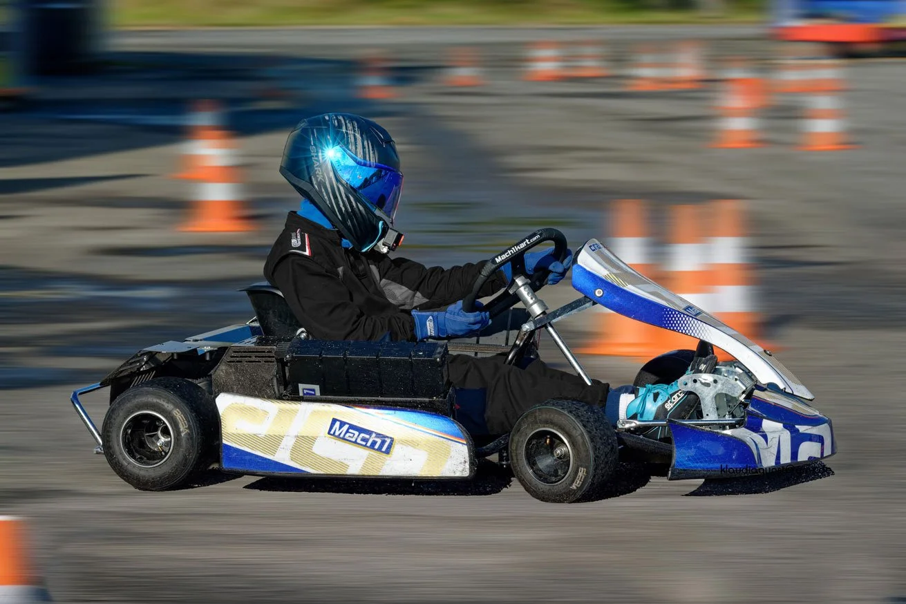 Ein Fahrer auf einem go-kart-artigen Fahrzeug bei einem Fahrtraining mit orangenen Verkehrsleitkegeln im Hintergrund, trägt einen Helm und eine Schutzkleidung