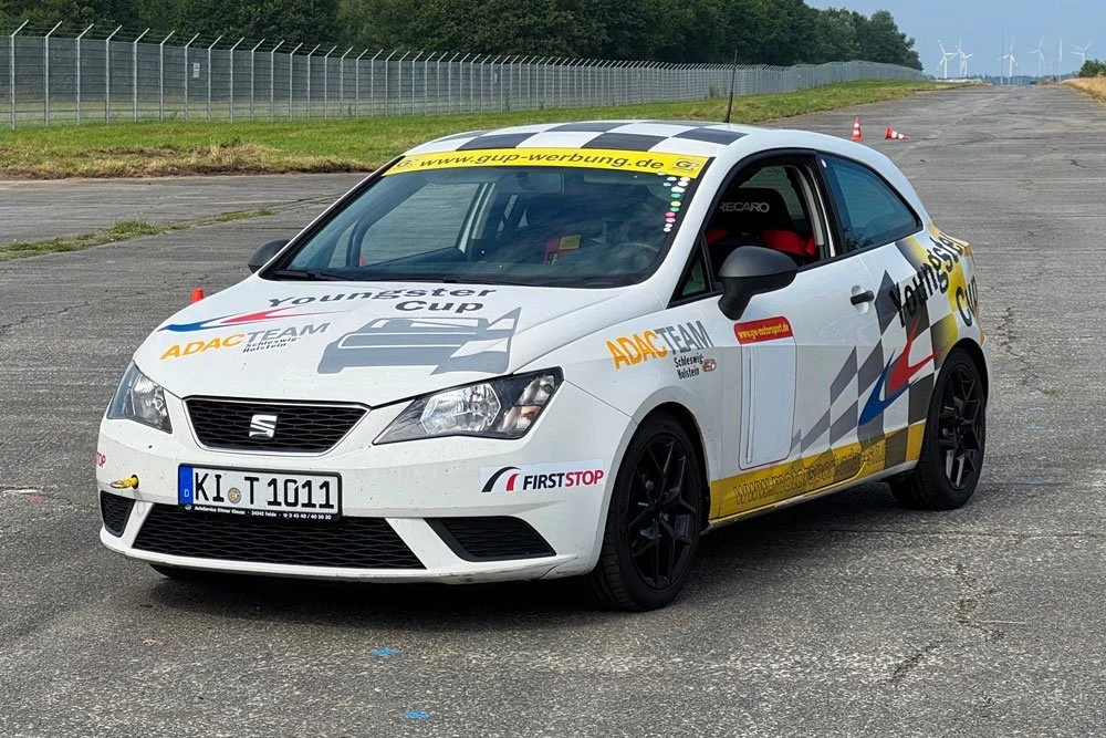 Ein weißes Rennauto mit ADAC, Youngster Cup und anderen Sponsorenaufklebern, steht auf einer Rennstrecke neben einem Zaun. Im Hintergrund sind Bäume und Windkraftanlagen zu sehen.